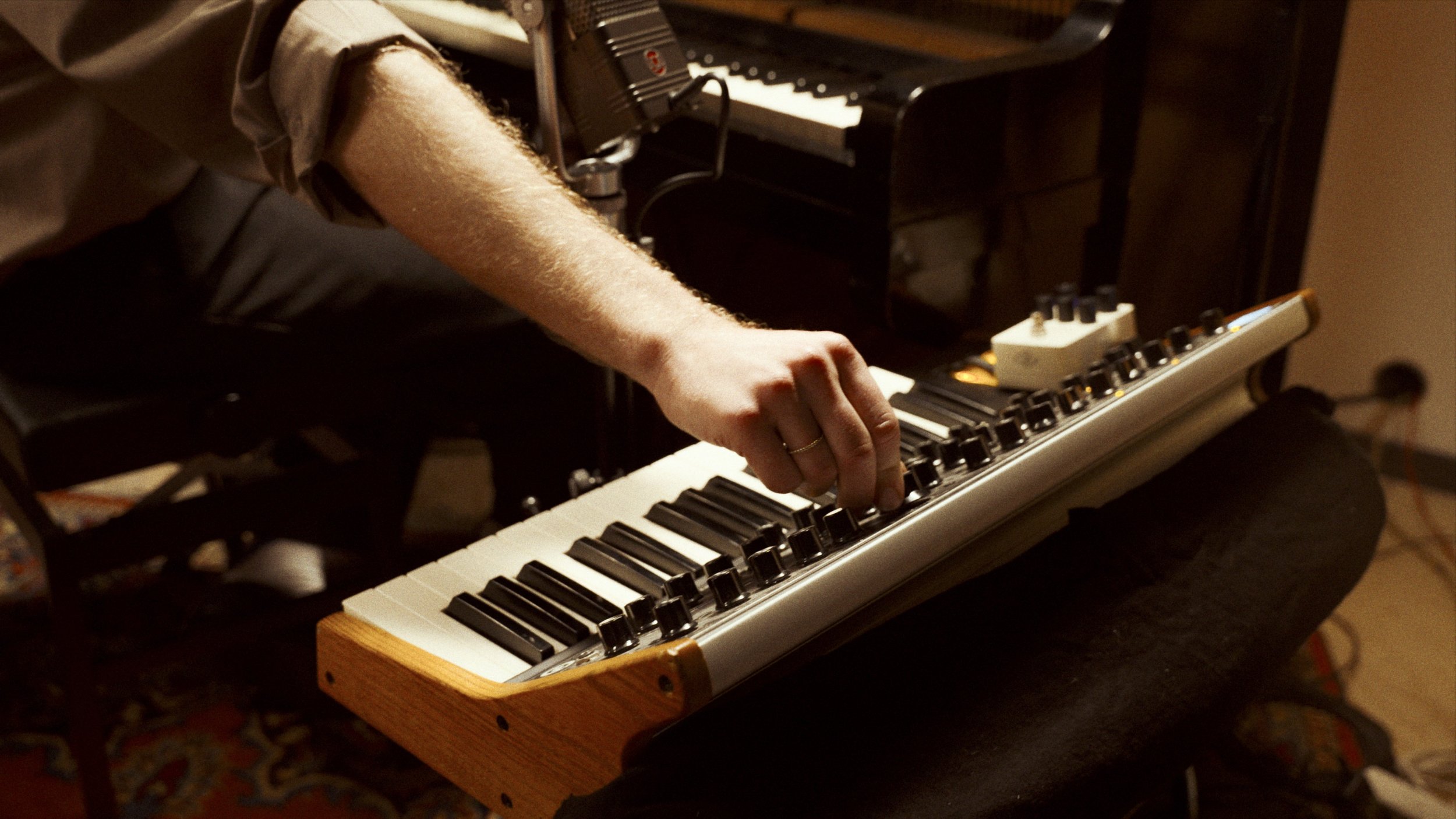 Close-up of a person's hand adjusting the knobs on a small electronic keyboard with a wooden frame, with an upright piano in the background.