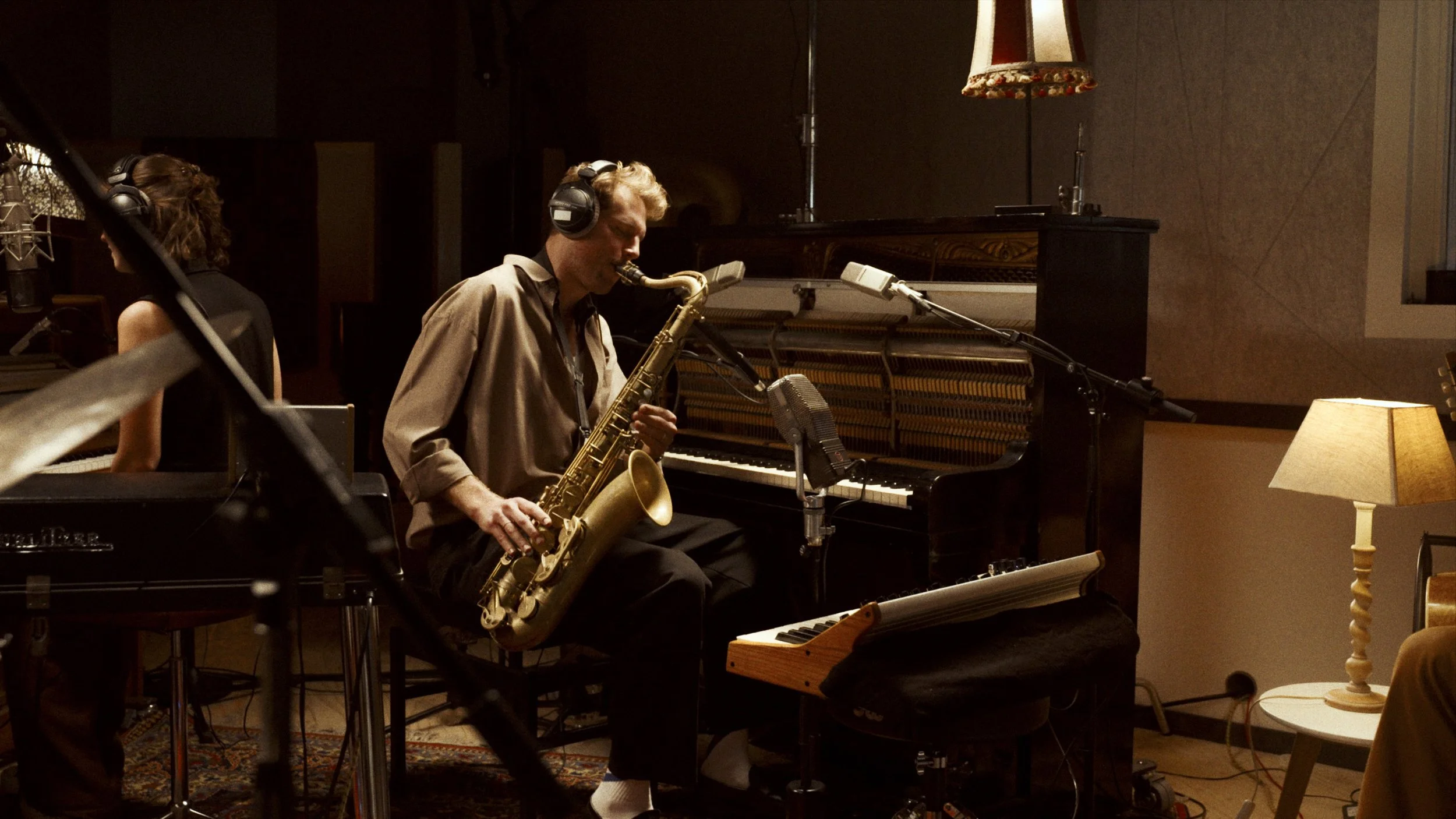 A man playing the saxophone in a recording studio with a woman at a piano nearby. The room has warm lighting and vintage decor, including a lamp on a side table.