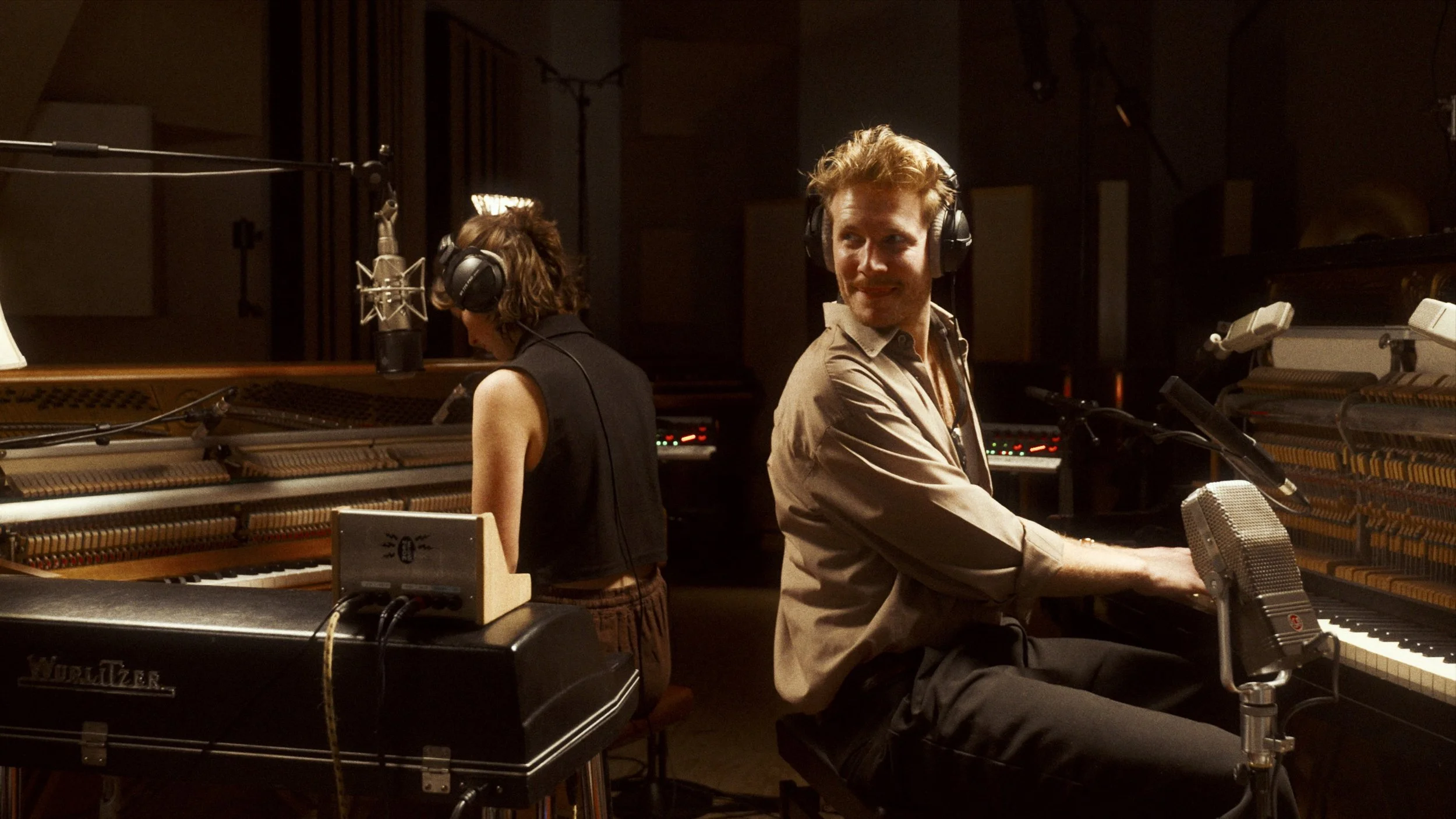 Two musicians in a recording studio, sitting at pianos with microphones, wearing headphones, one woman with short hair and one man with red hair, both focused on their instruments.