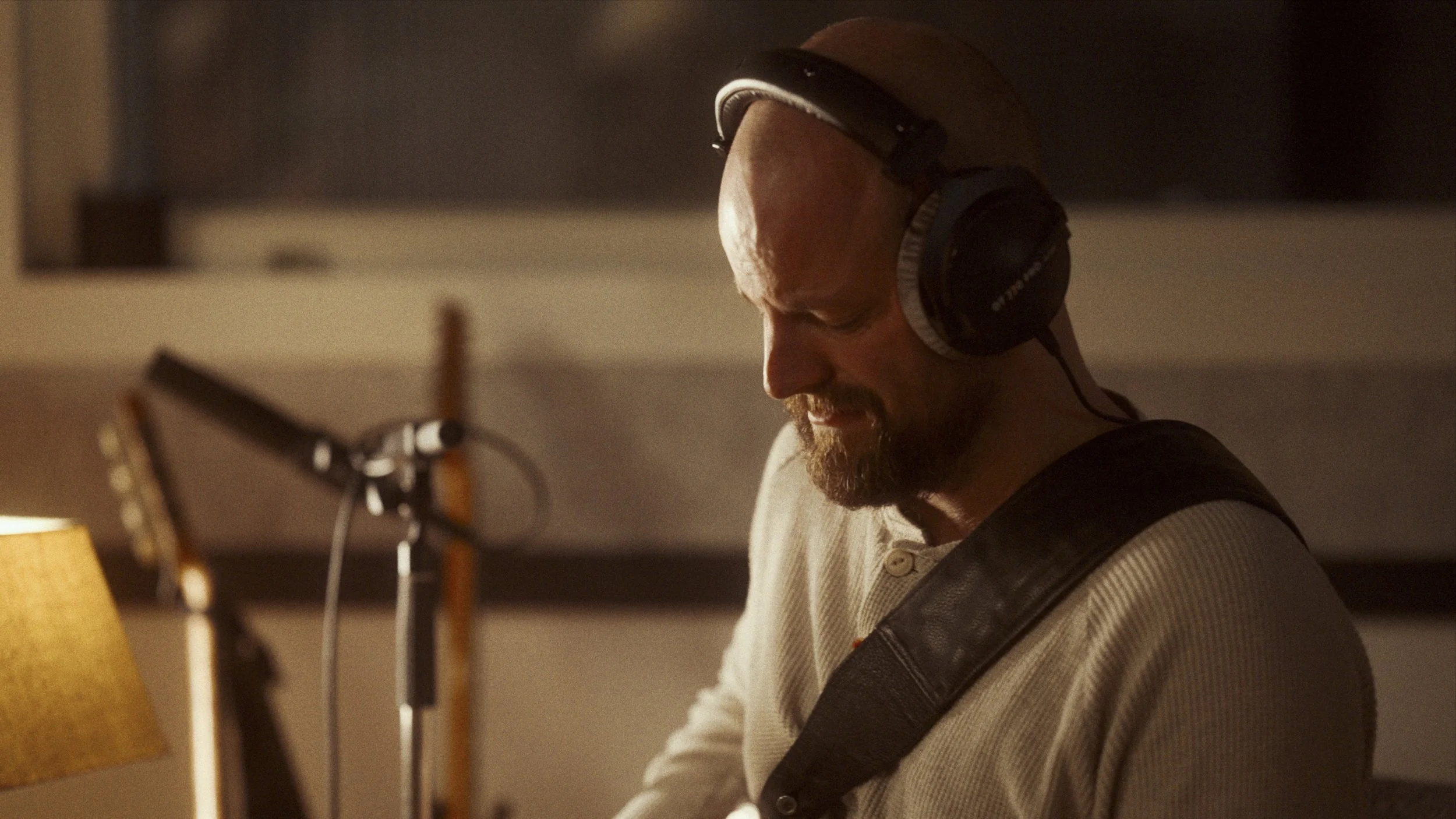 Man with a beard wearing headphones and a beige shirt playing guitar in a recording studio.