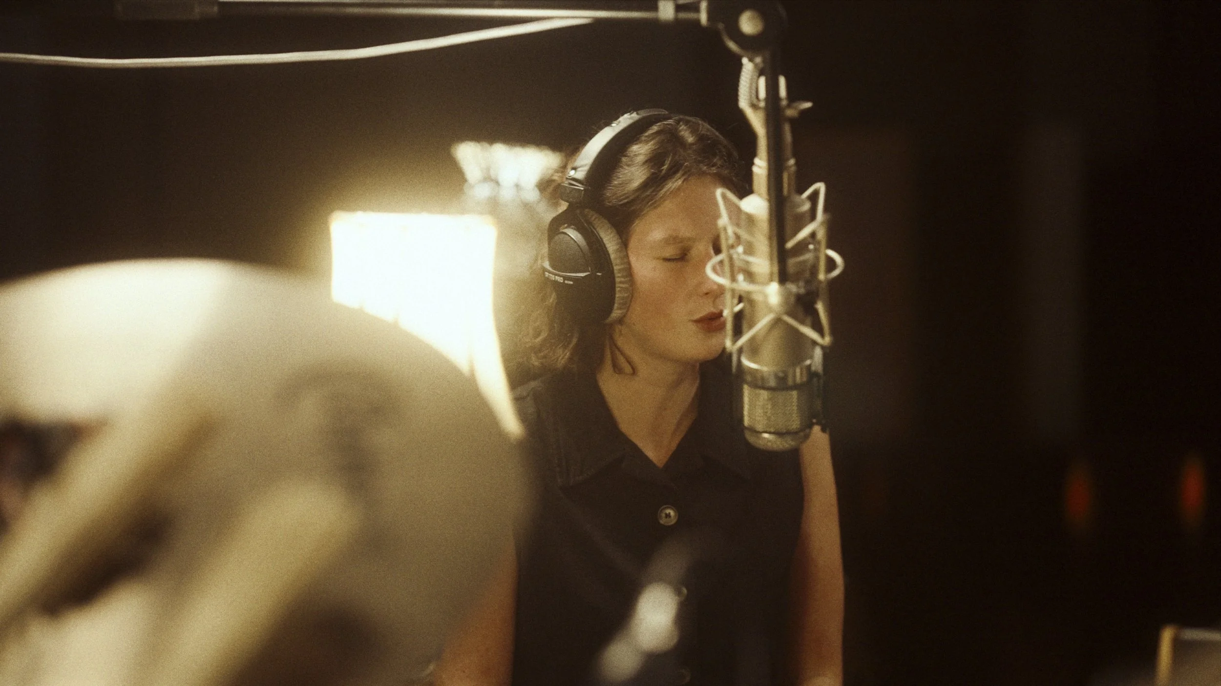 A woman with short brown hair wearing headphones, singing into a professional microphone in a recording studio with dim lighting.