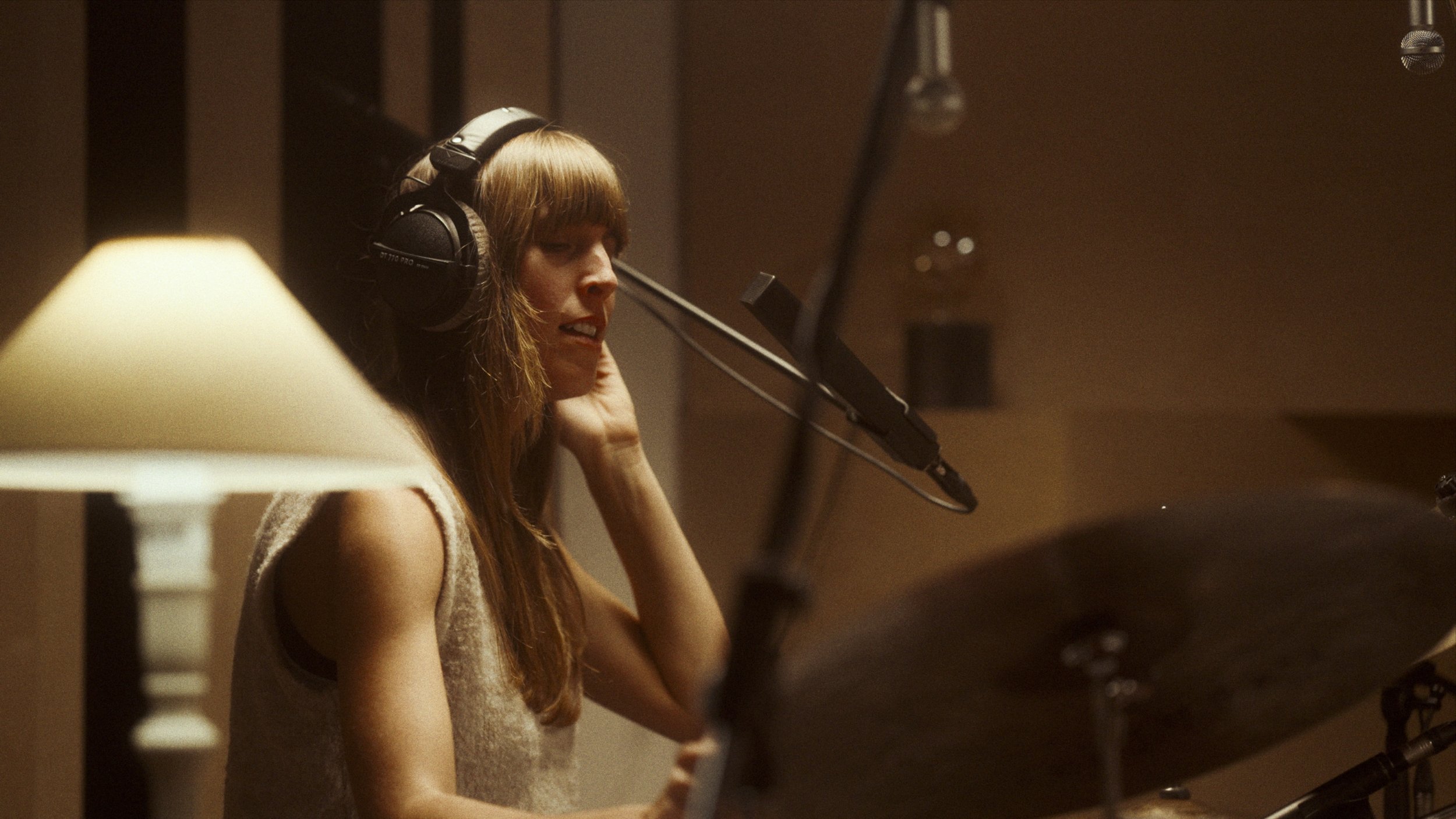 A woman with long hair in a recording studio, wearing headphones, singing into a microphone with a large cymbal in the foreground.
