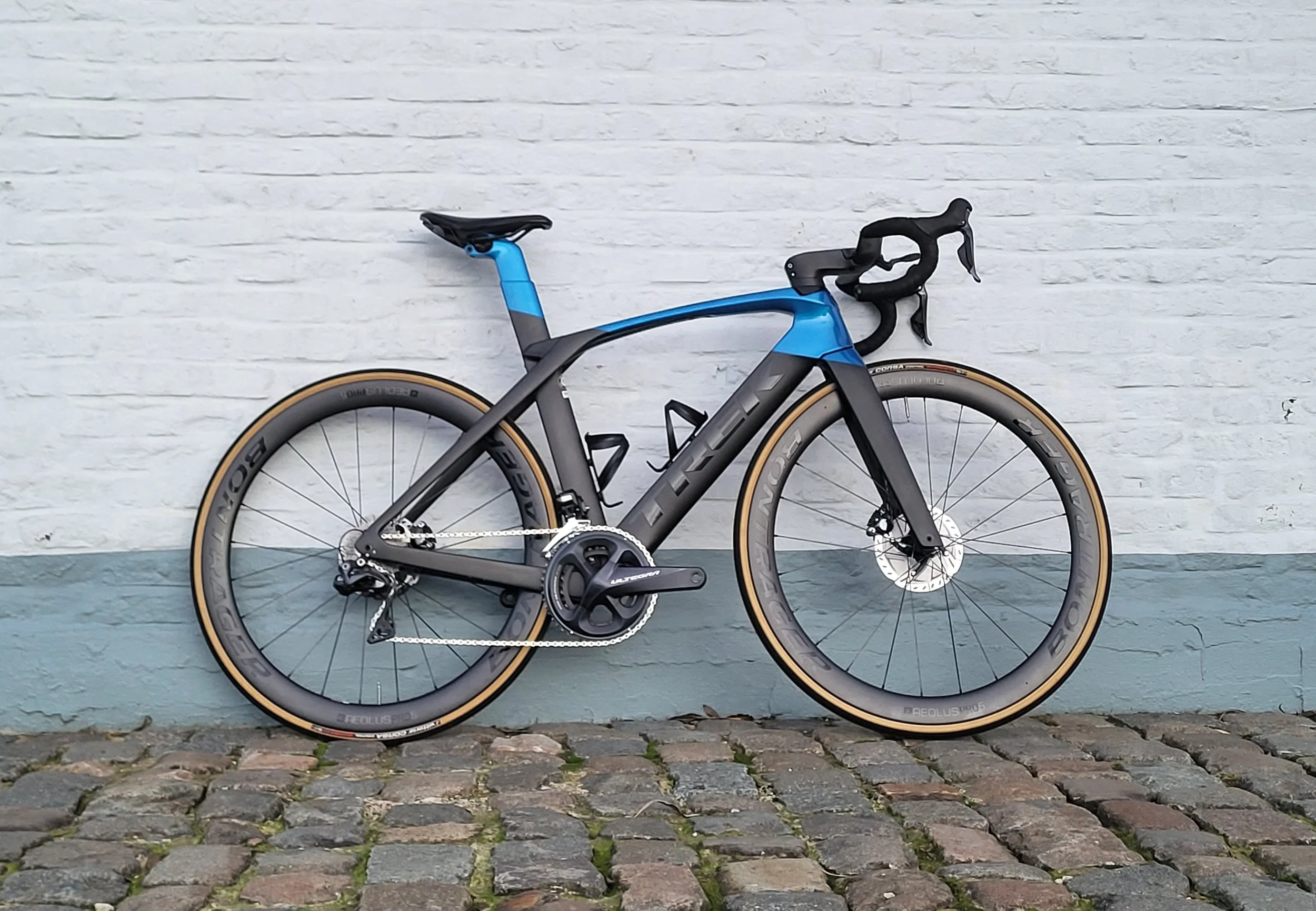 Een zwart en blauw racefiets met dunne banden, liggende op een stenen straat voor een witte bakstenen muur.