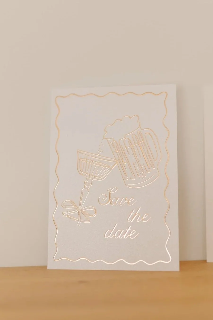 Een witte kaart met gouden lijnen en tekst dat zegt 'Save the date', versierd met een beeld van een bierglas en een schaal met een toost.