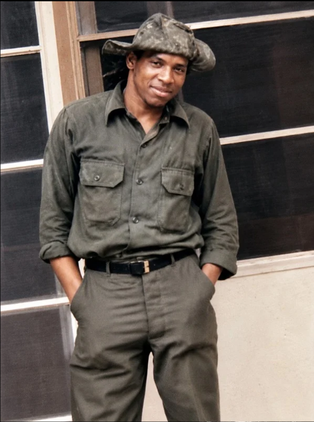 A woman dressed in a military-style uniform with a matching hat, standing indoors with her hands in her pockets, smiling slightly.