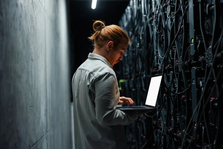 A woman in a beige blazer and white shirt holding a tablet, standing in a data center with purple and blue lighting.