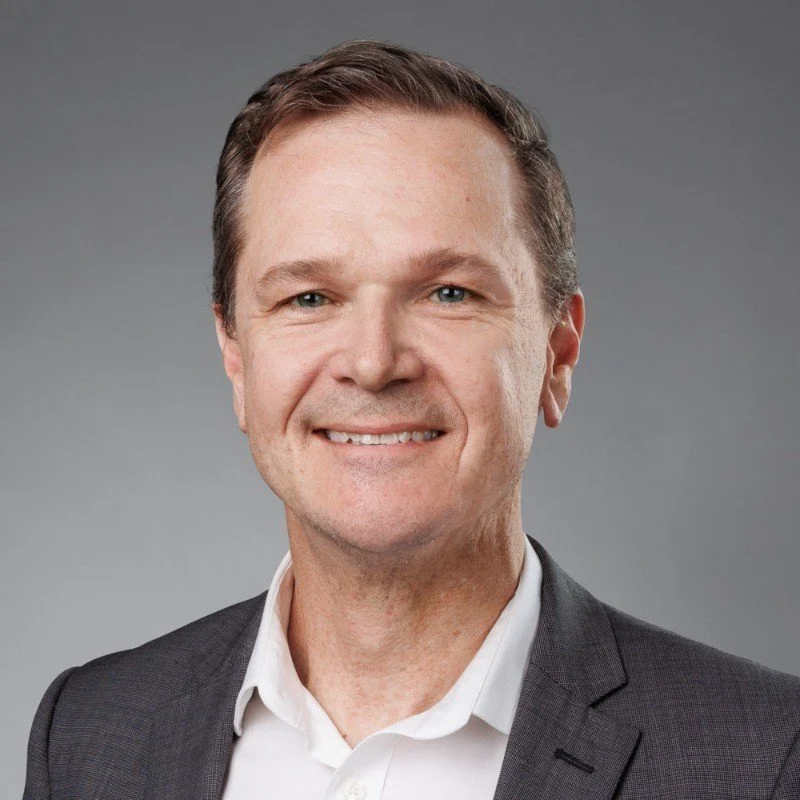 A middle-aged man with short brown hair, wearing a dark blazer over a white collared shirt, smiling against a plain gray background.