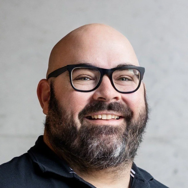 A smiling man with a beard, glasses, and a bald head, wearing a black jacket against a light background.