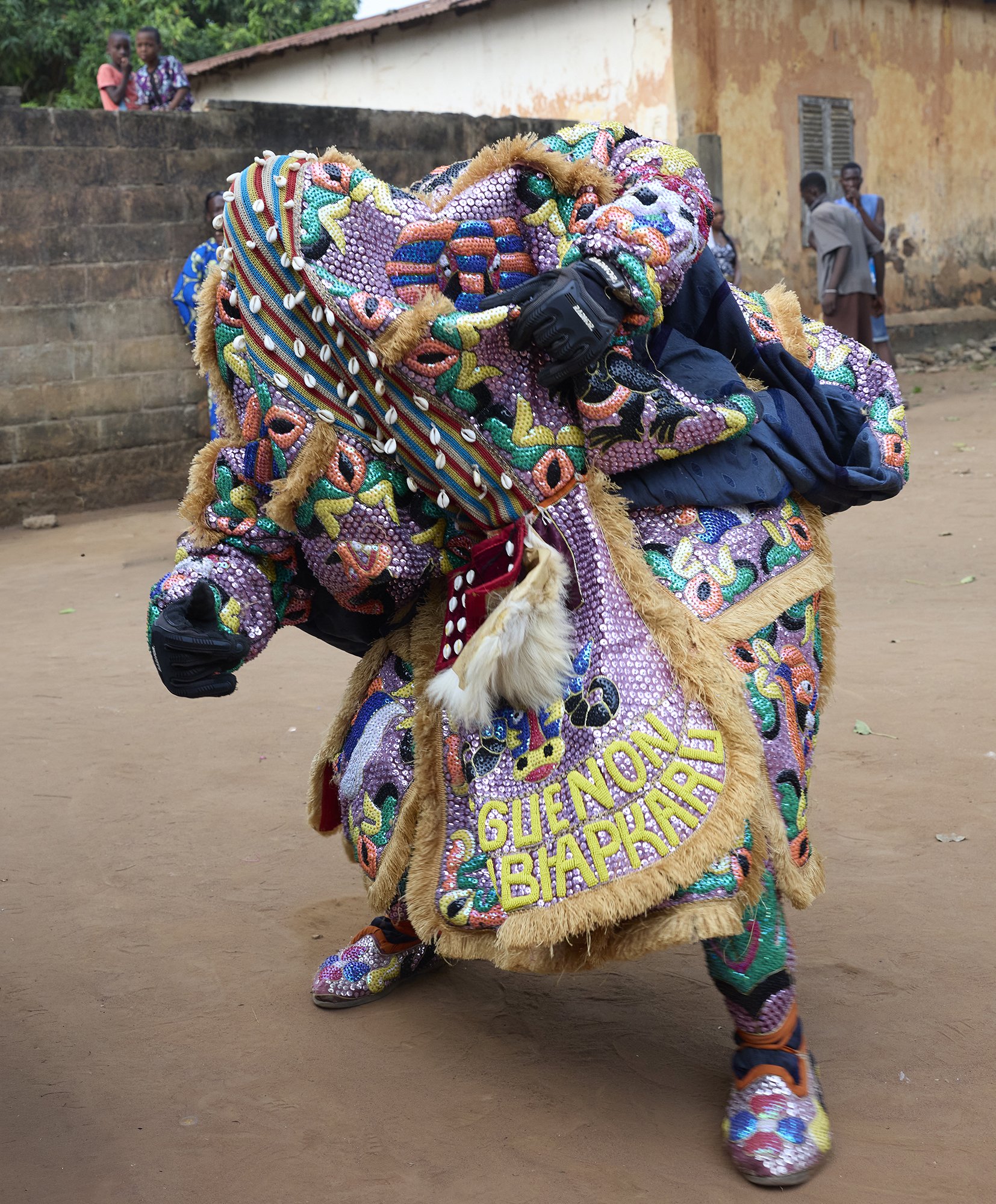 Une personne en costume traditionnel coloré avec des paillettes et des motifs apparaît dans cette image, marchant ou dansant avec des enfants et des adultes en arrière-plan dans une zone extérieure avec un bâtiment en pierre et un sol en terre.