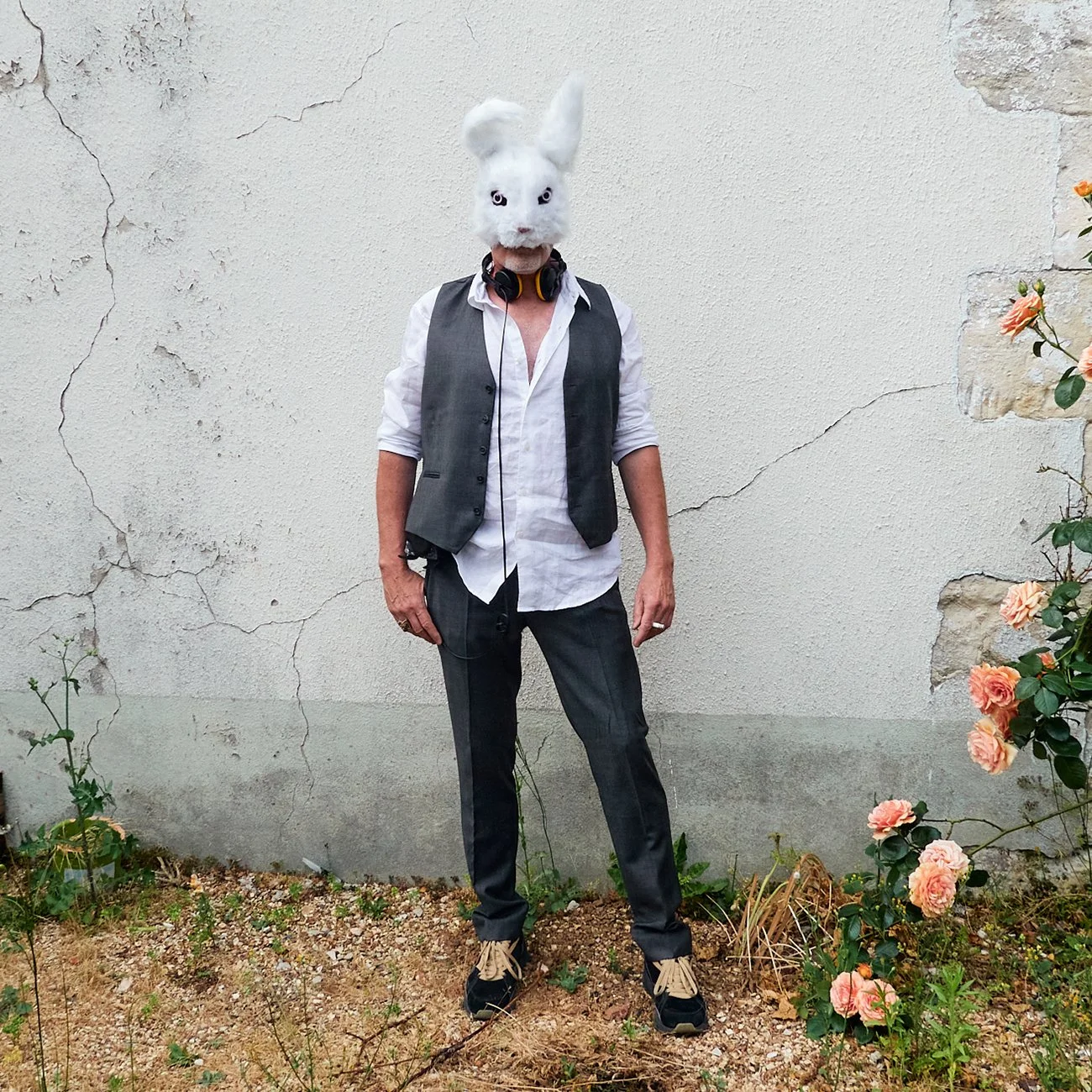 Personne portant un masque de lapin blanc et des écouteurs, vêtue d'une veste grise, chemise blanche, pantalon gris, debout devant un mur pâle avec des plantes à droite.