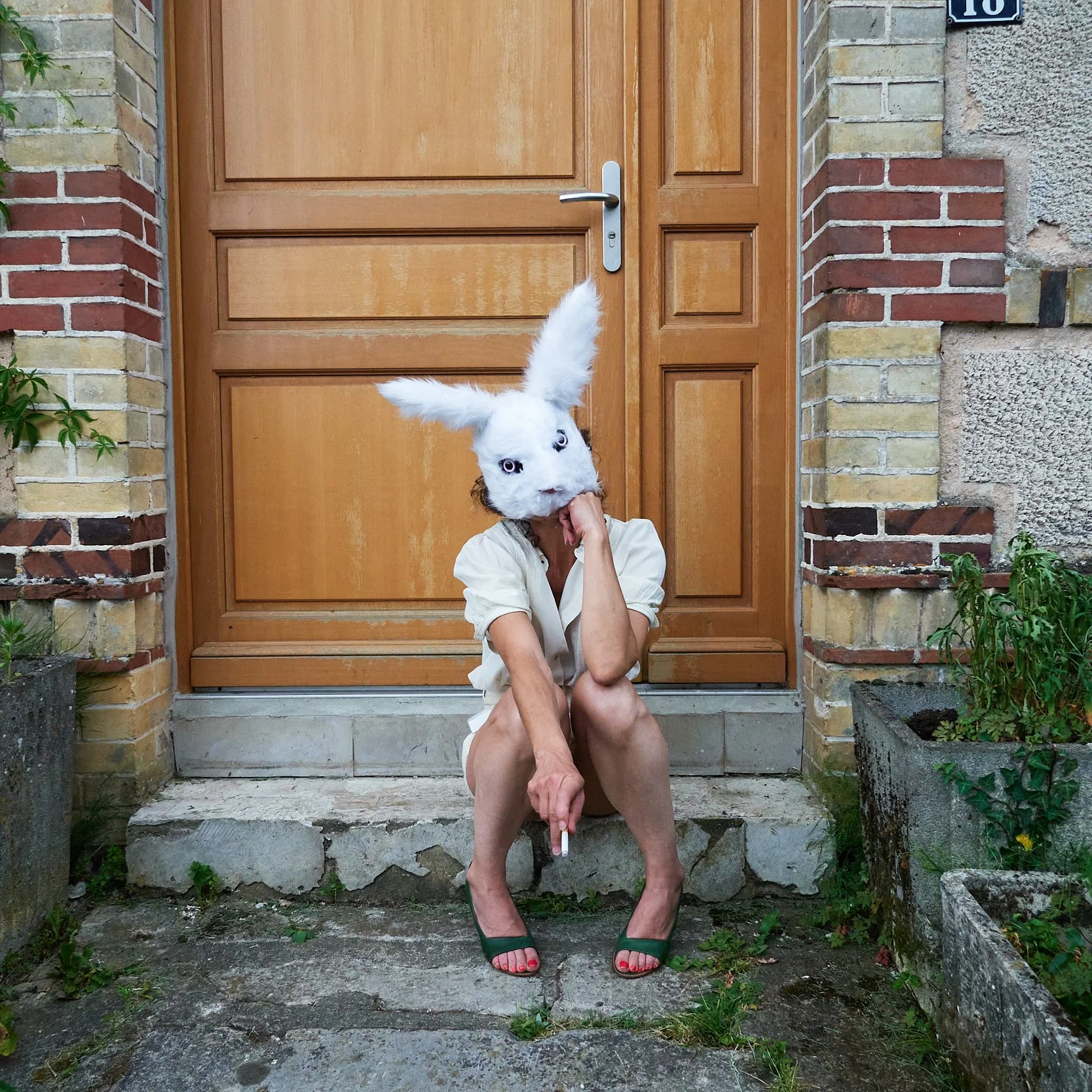 Personne assise devant une porte en train de fumer, portant un masque de lapin blanc, robe blanche, chaussures vertes, les cheveux courts.