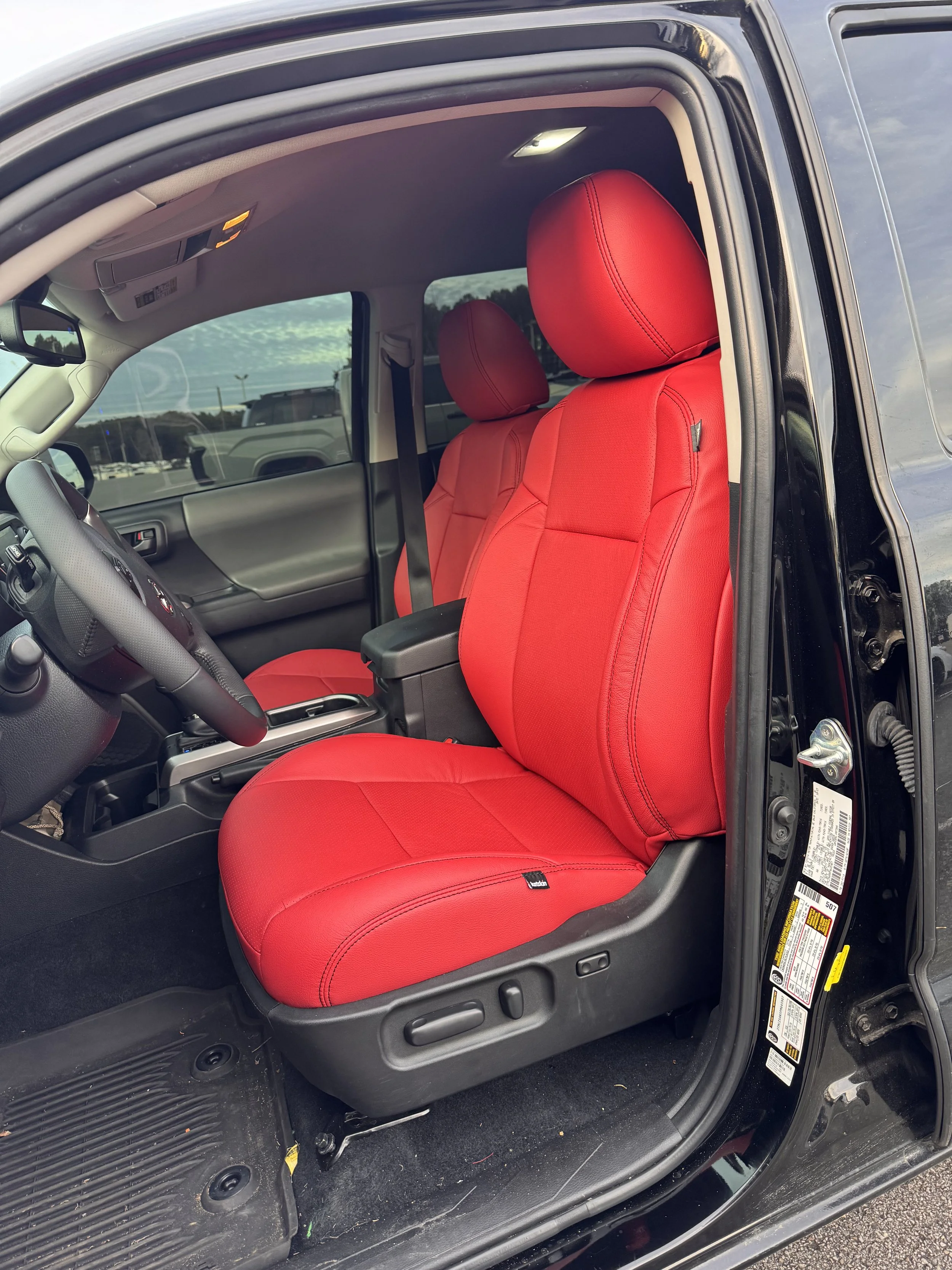 The front interior of a black vehicle with red seat covers on the driver and passenger seats, a black dashboard, and a black floor mat.