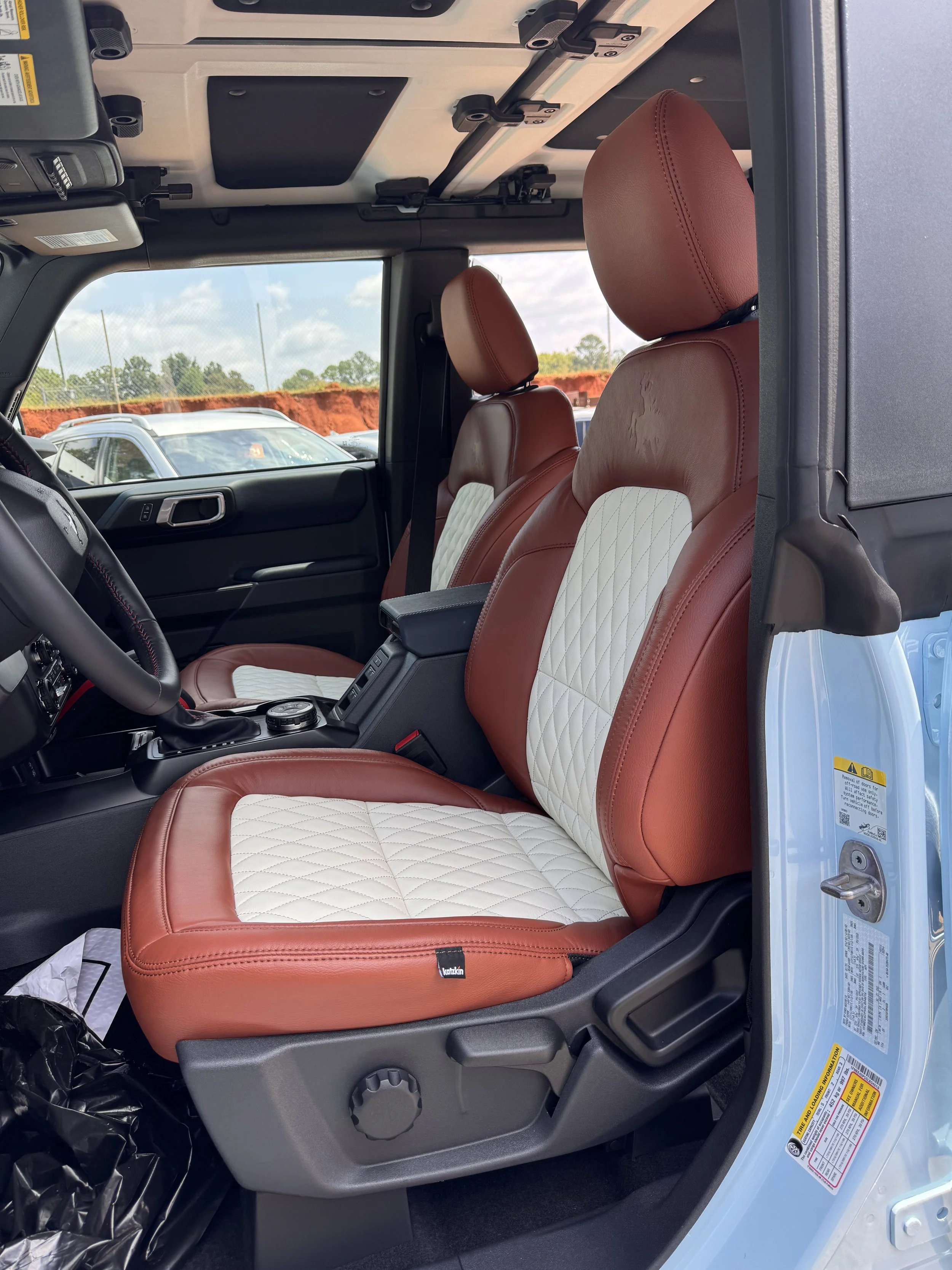 Interior of a vehicle with red and white quilted leather seats, black dashboard, and windows showing outdoor parking lot and cloudy sky
