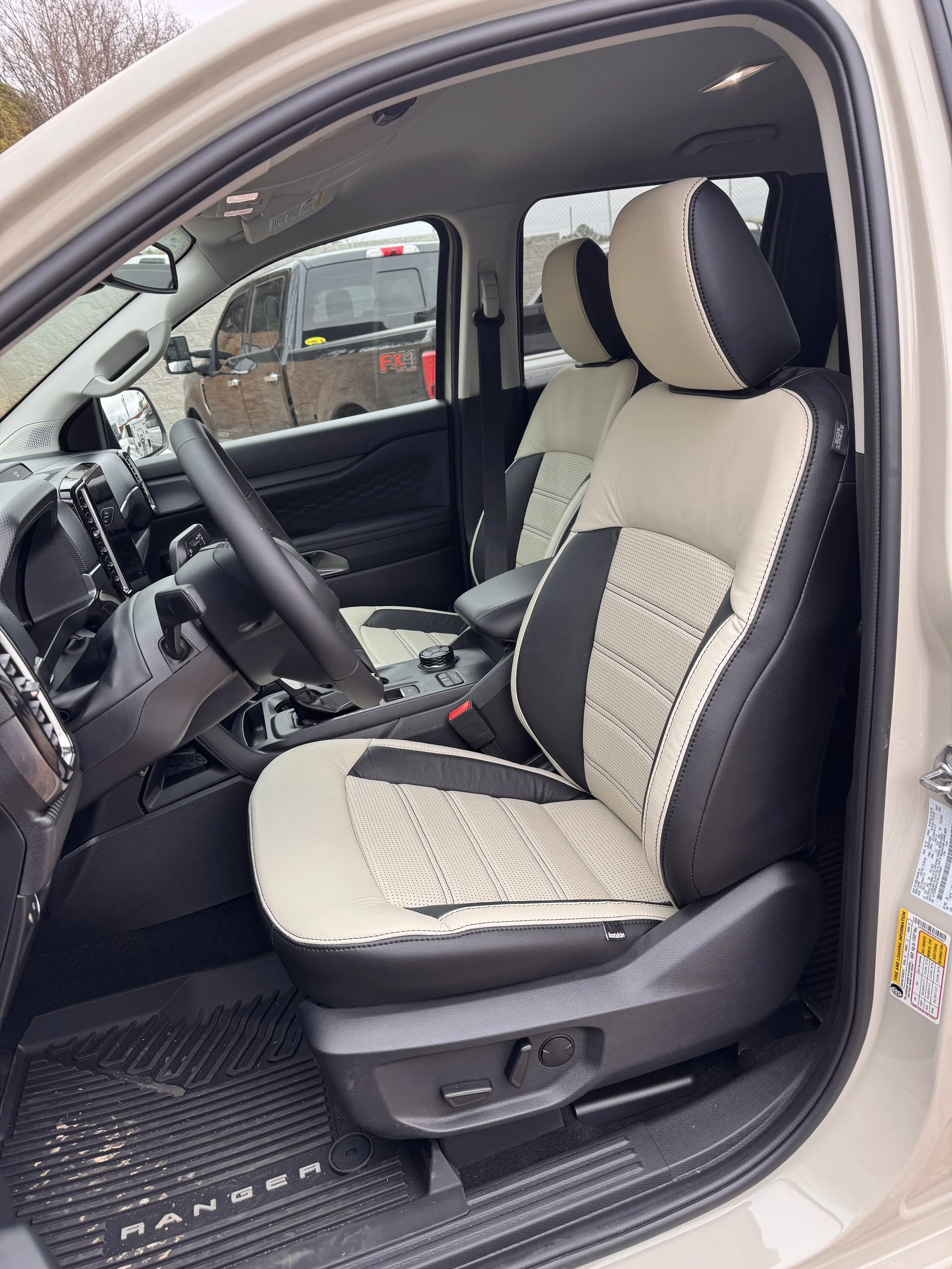 Front interior of a beige and black Ford Ranger pickup truck showing driver's and passenger's seats, steering wheel, dashboard, and part of the center console, with a view of other trucks through the window.