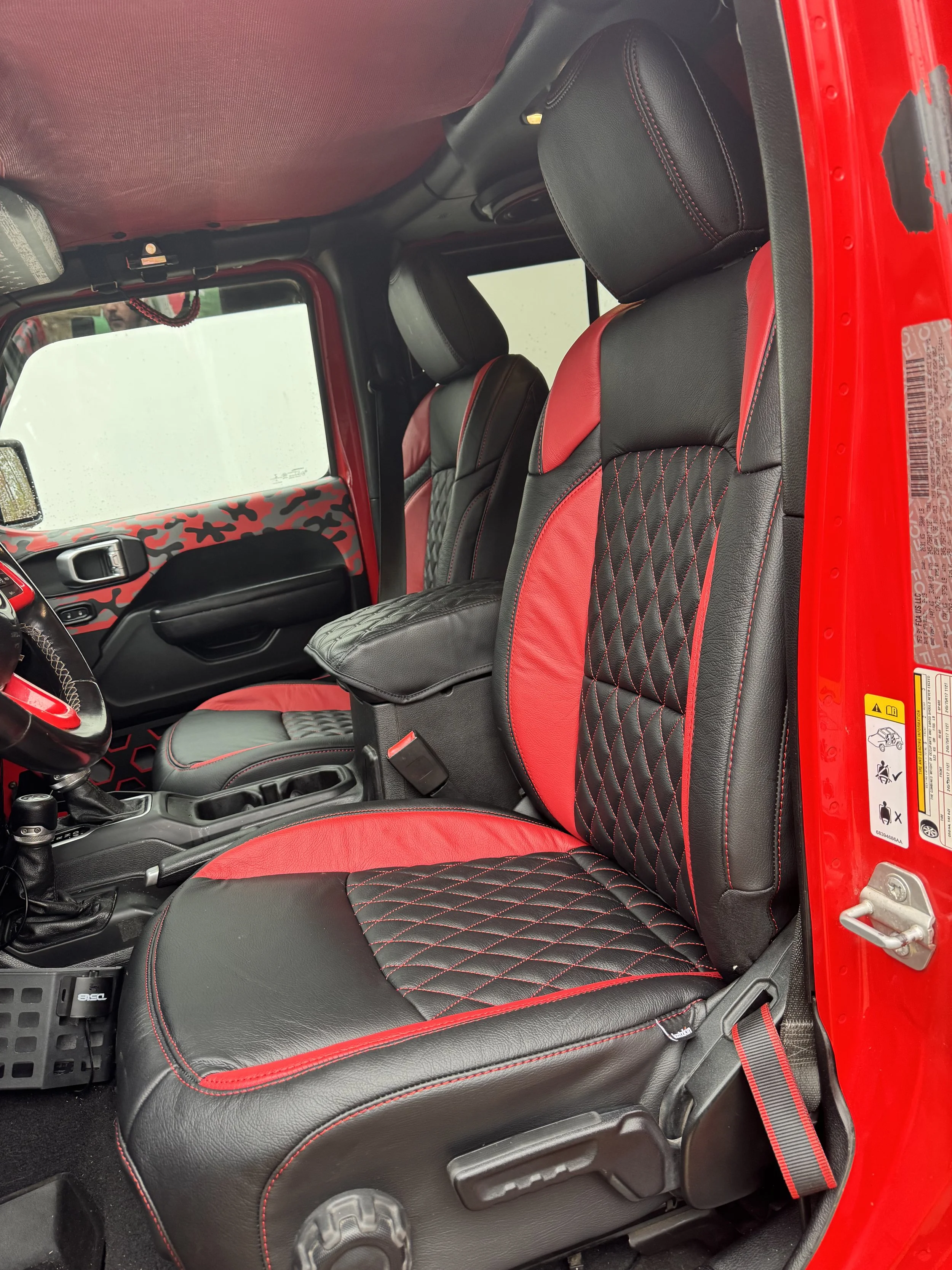 The interior of a red vehicle showing black leather seats with red accents and diamond stitching, along with a red and black camouflage pattern on the dashboard and door panel.