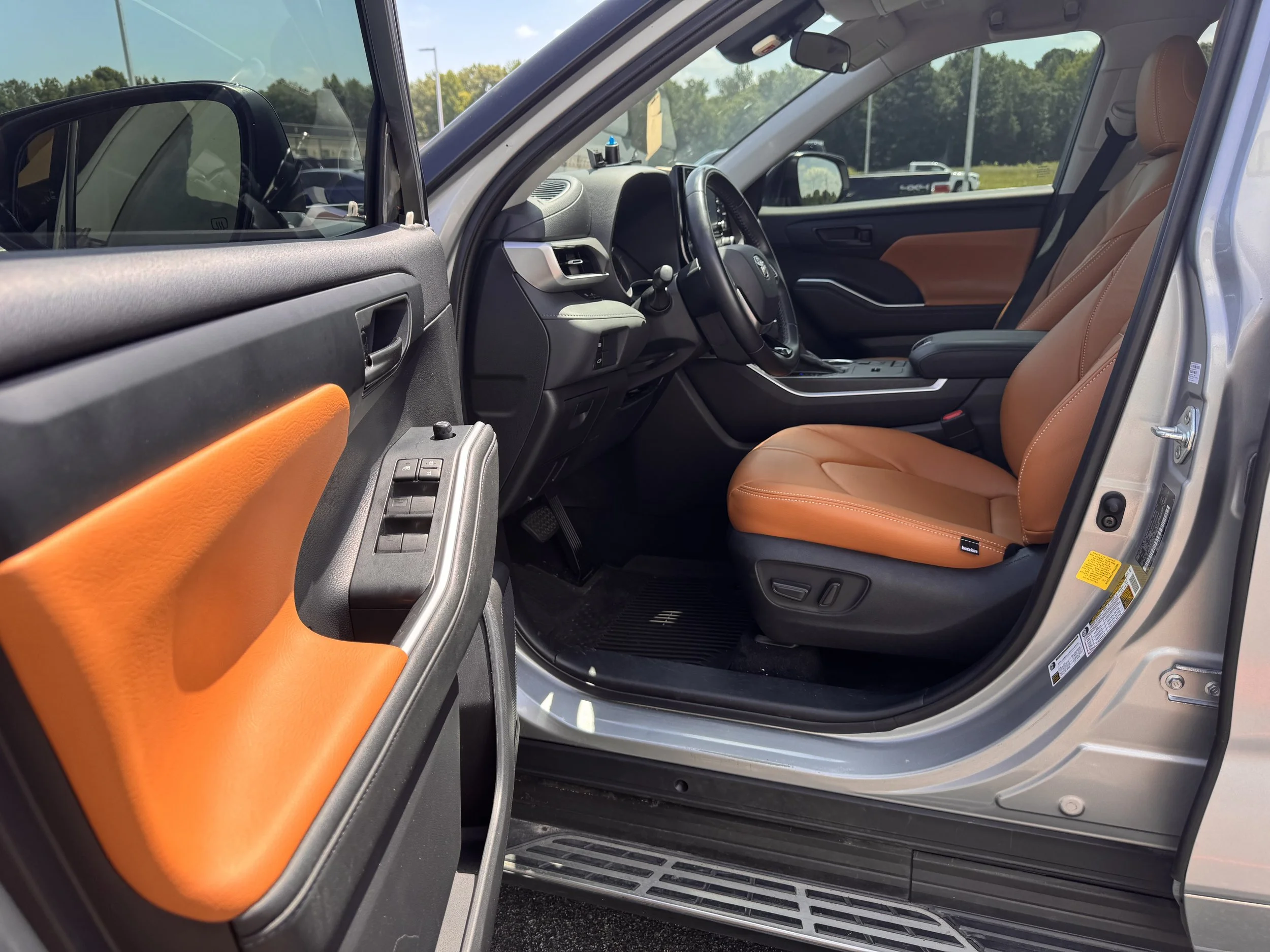 Inside view of a vehicle's front passenger side, showing the dashboard, brown leather seat, black interior with orange accents on the door panel, and a glimpse of the outside parking lot with other vehicles and trees.