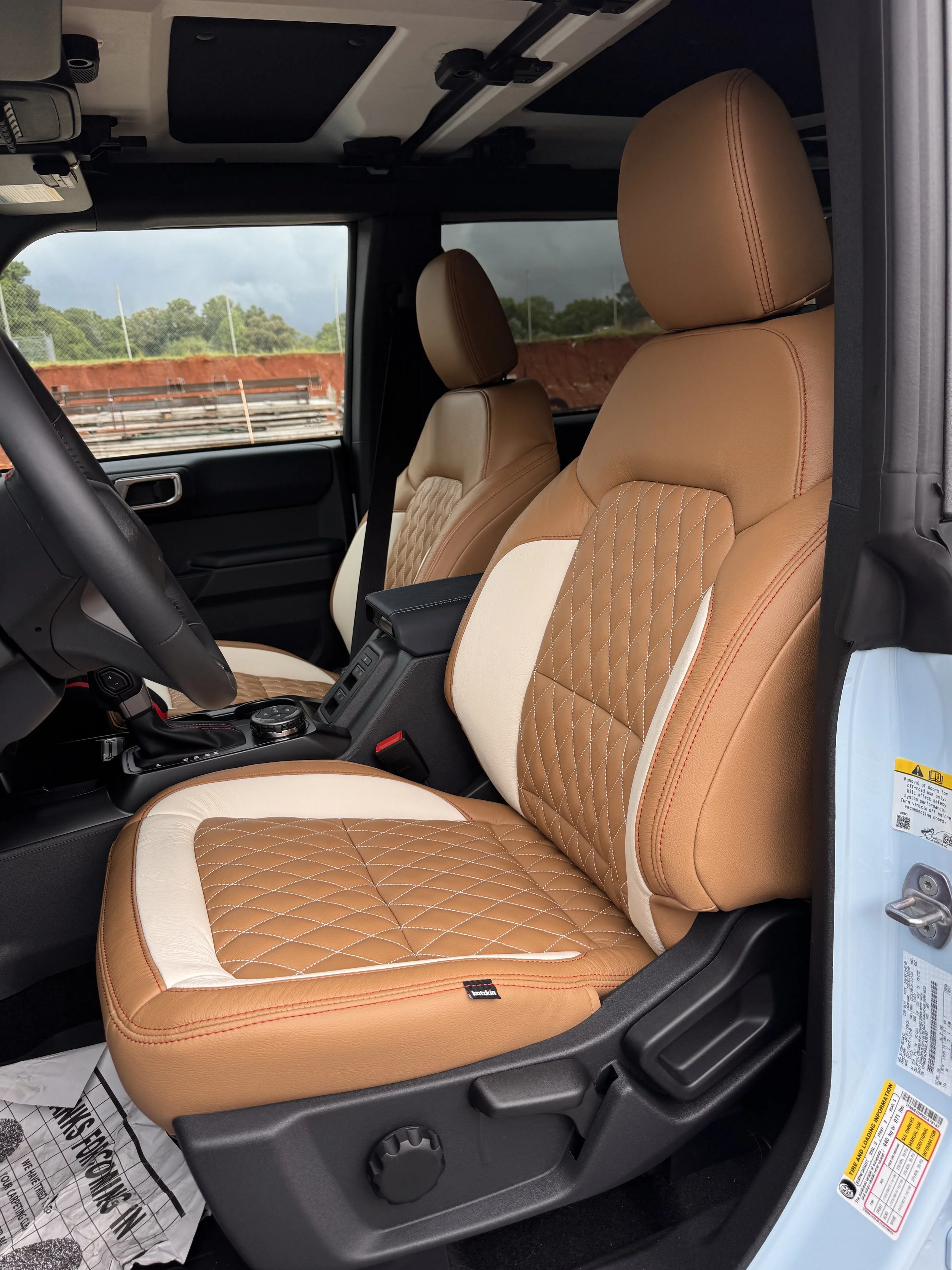 Interior of a vehicle showing tan leather seats with diamond stitching, with a black dashboard and window showing an outdoor scene with trees and cloudy sky.
