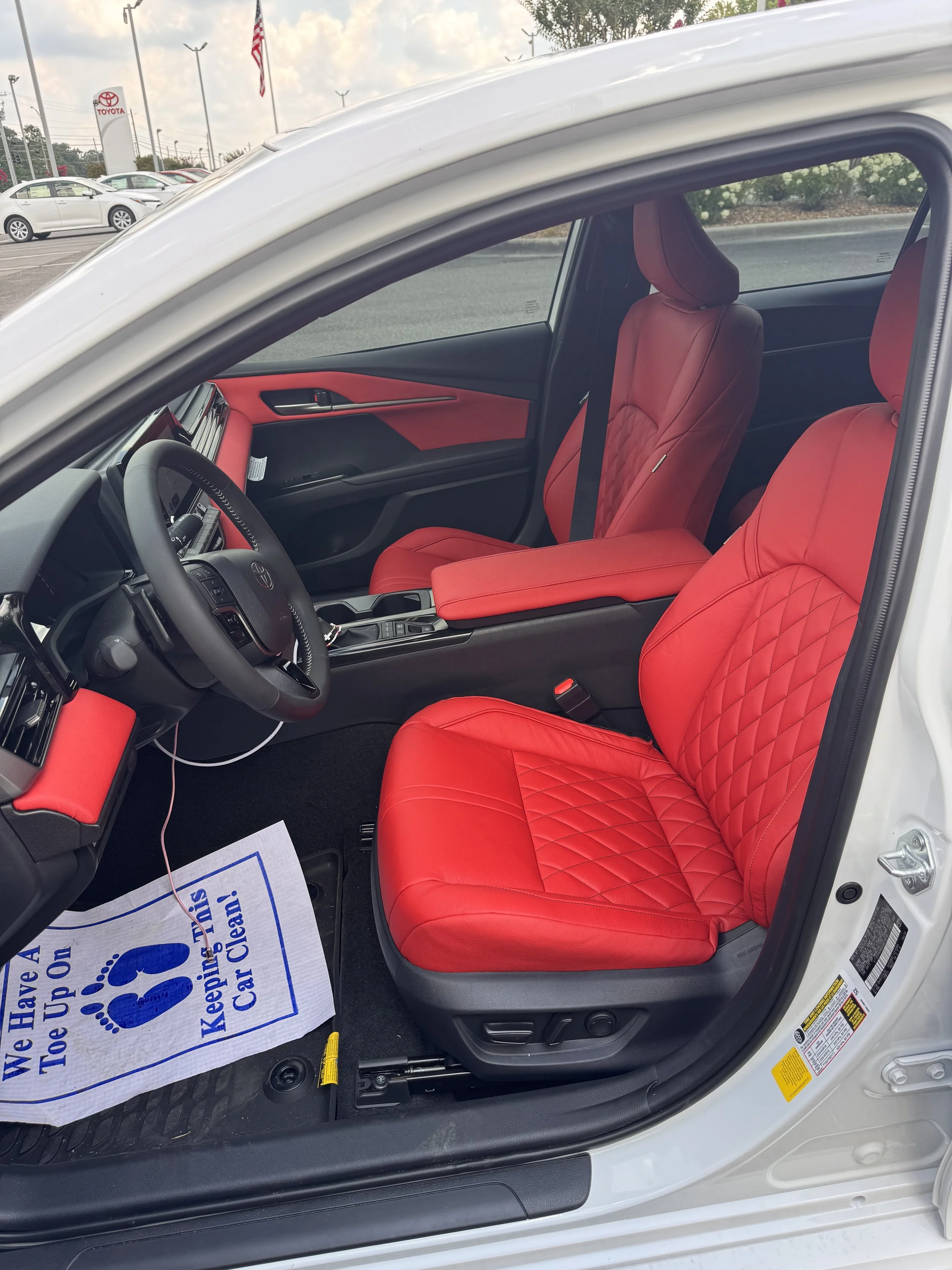 Interior of a white car with red quilted seat covers, black dashboard, steering wheel, center console, and a gray floor mat with a sign that reads 'We Have A Toe Up On! Keep This Car Clean!'.