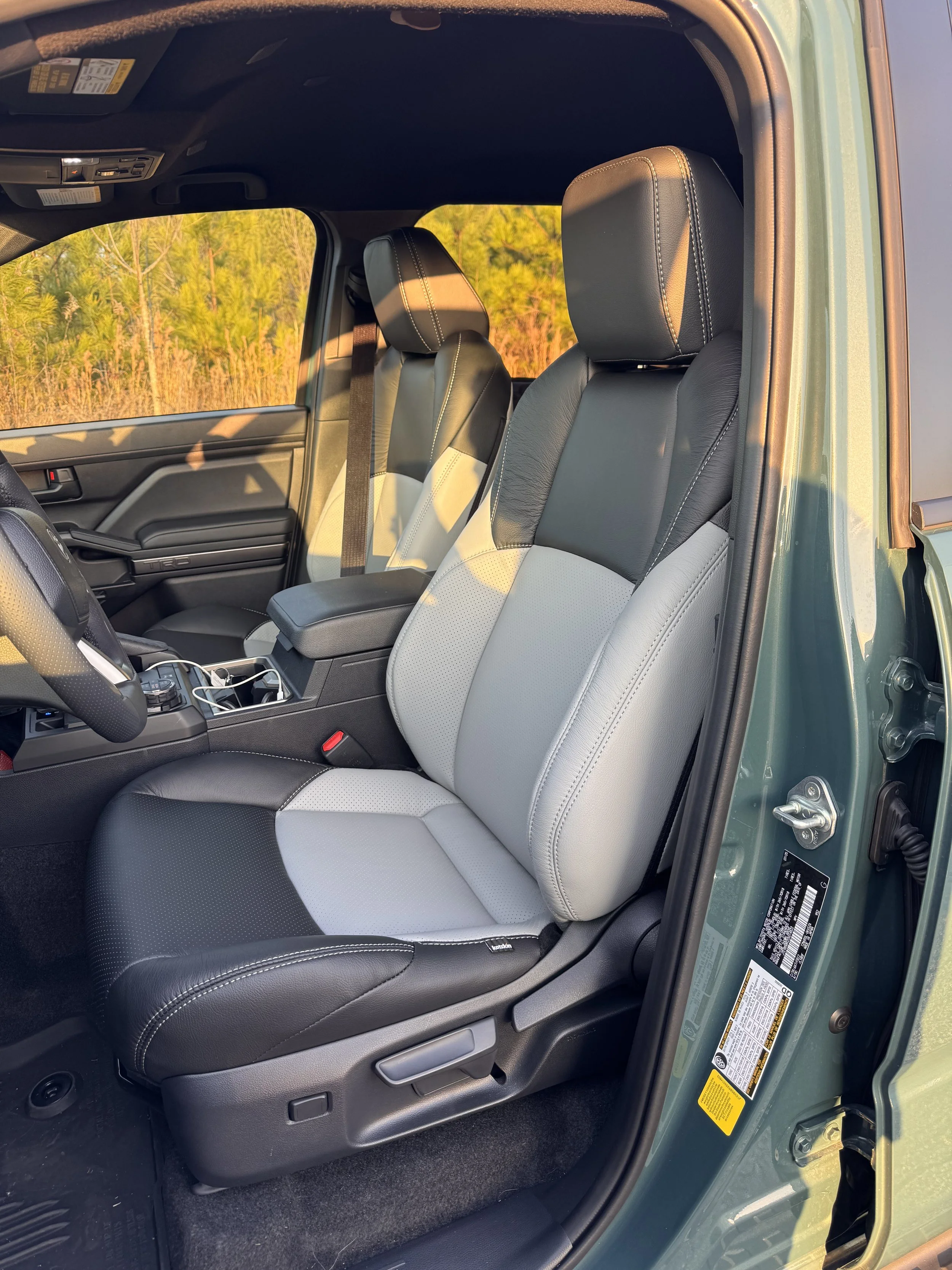 Inside view of a vehicle with black and light gray front seats, black dashboard, and open driver side door, with a background of trees and sunset lighting.