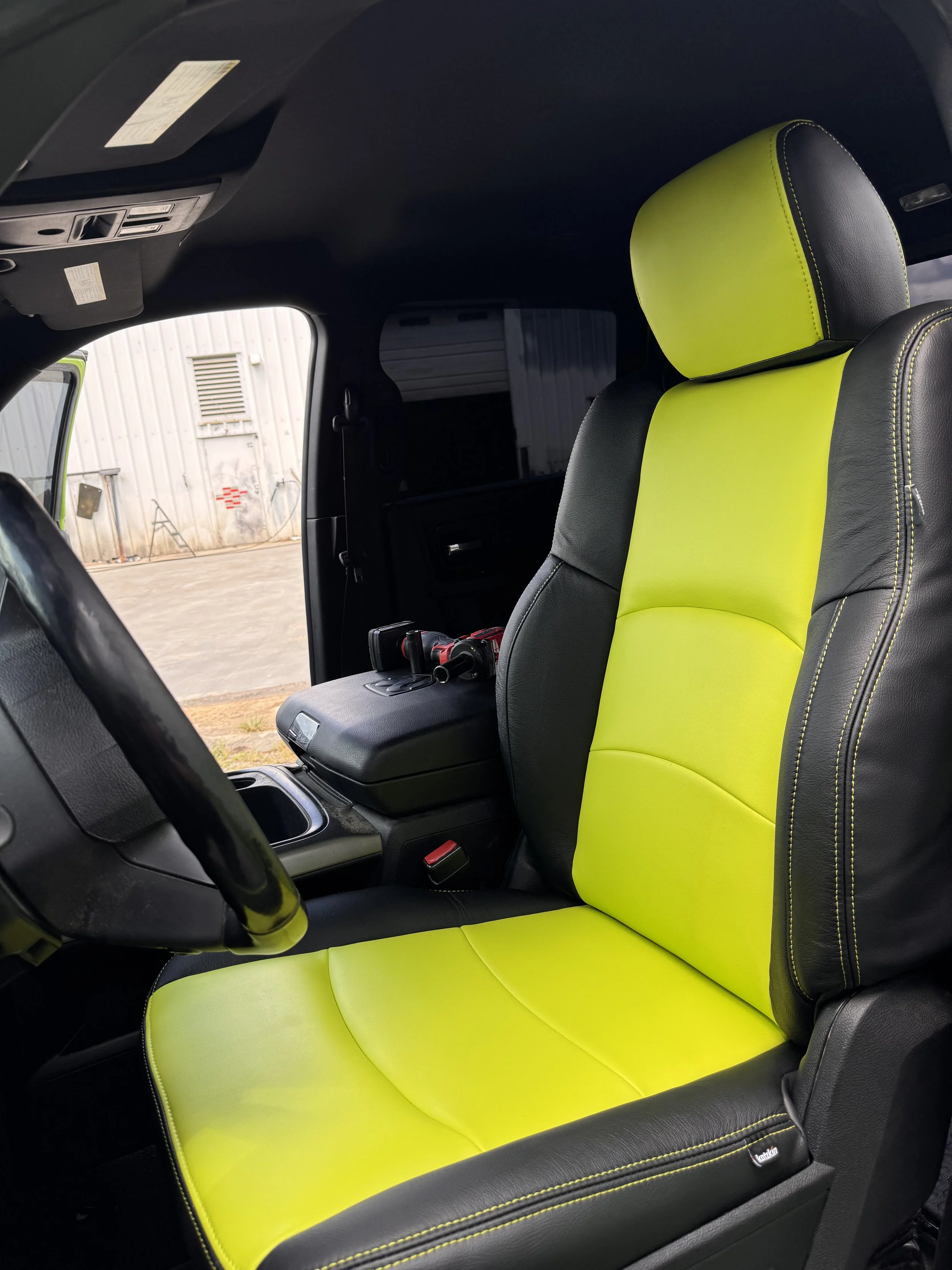 Interior of a vehicle showing a black and neon yellow seat cover on the driver's seat, with some tools on the center console and a view of a warehouse or garage through the window.