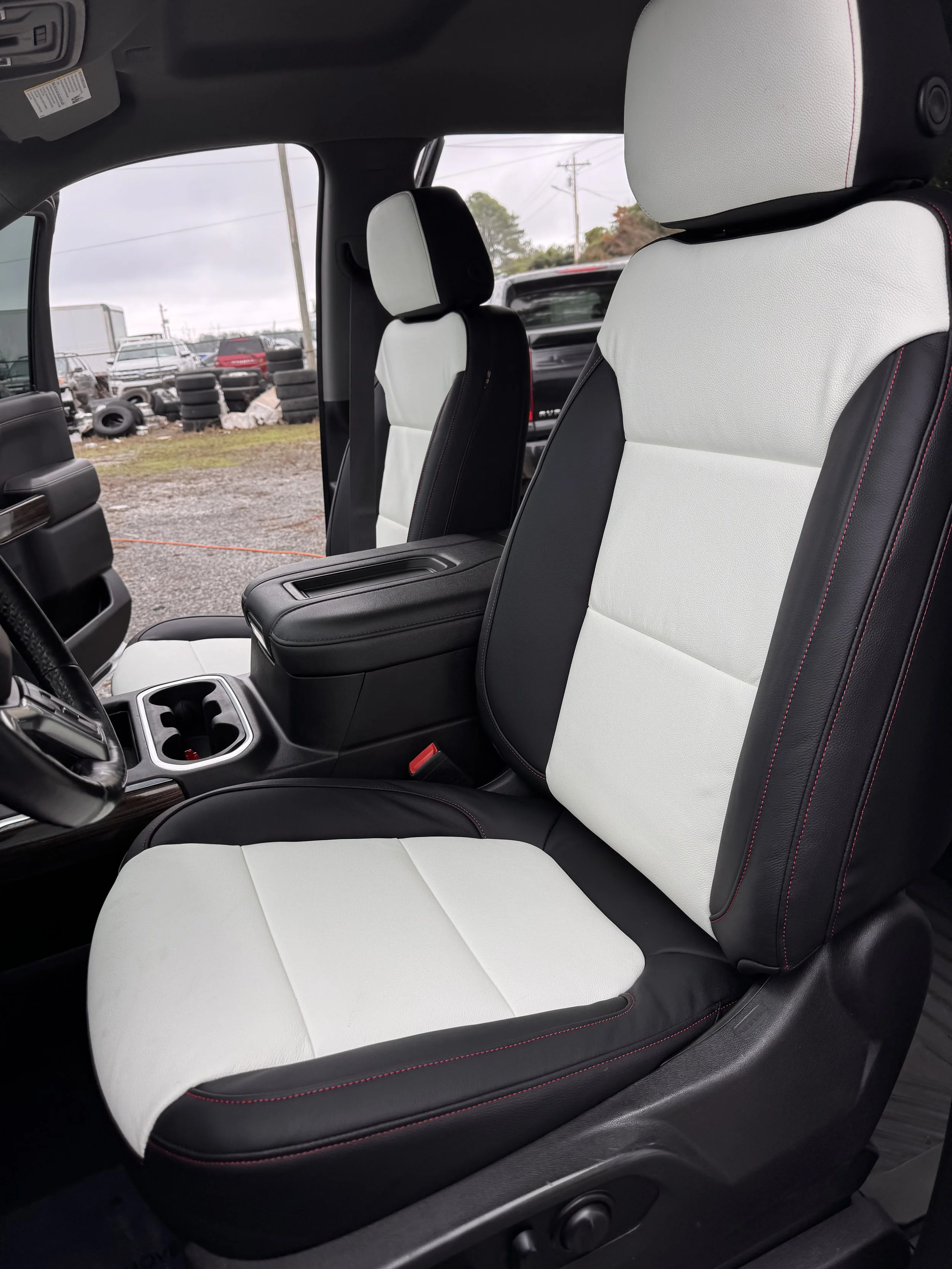 Inside view of a vehicle showing black and white front seats with red stitch detailing, center console with cup holders, and scenery of a tire yard outside.