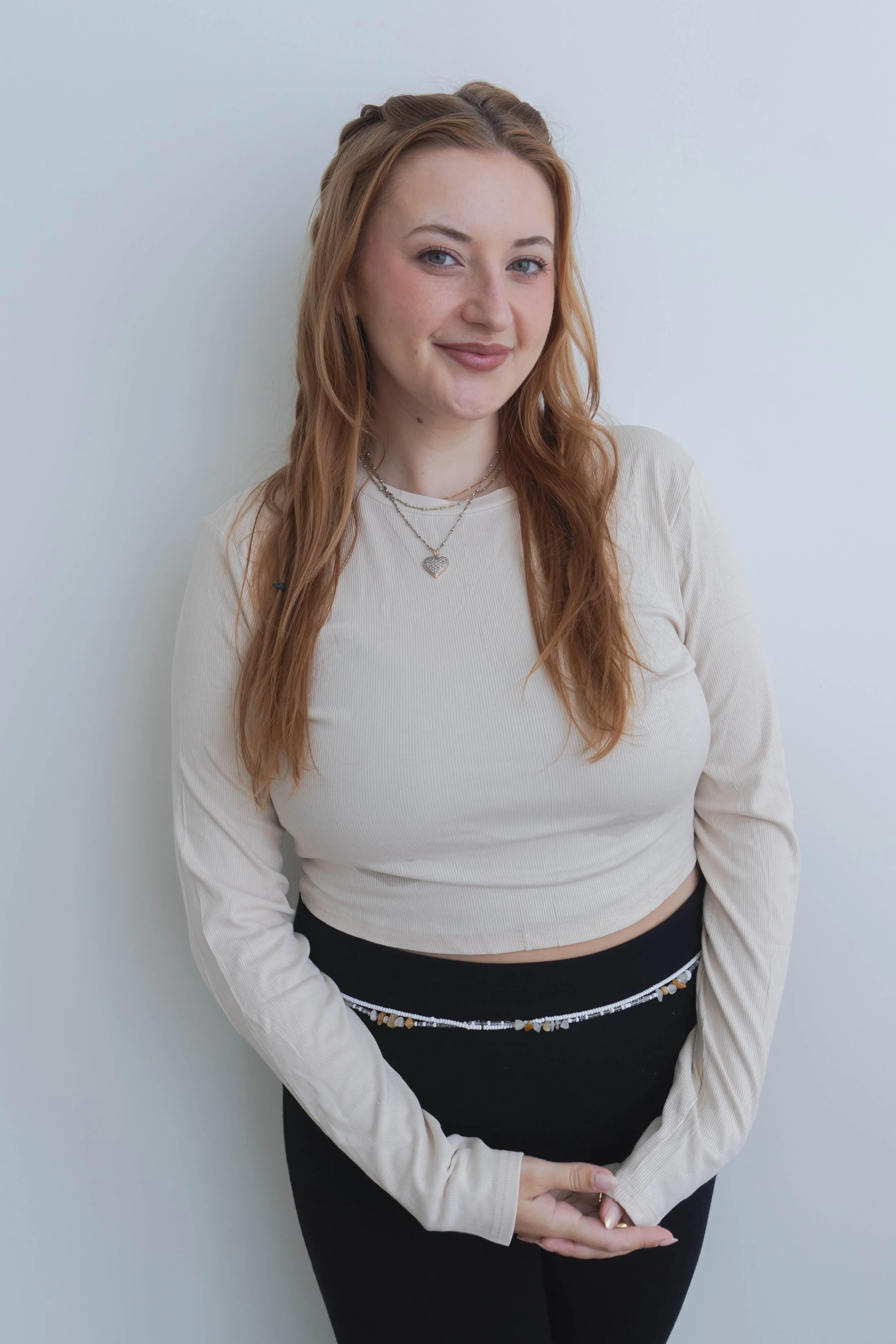 Young woman with long red hair, smiling, wearing a cream-colored long sleeve crop top, black pants, and layered necklaces, standing against a light gray wall.