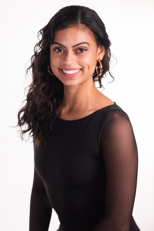 A woman with long curly dark hair, smiling, wearing a black sheer long-sleeve top and hoop earrings, posing against a white background.