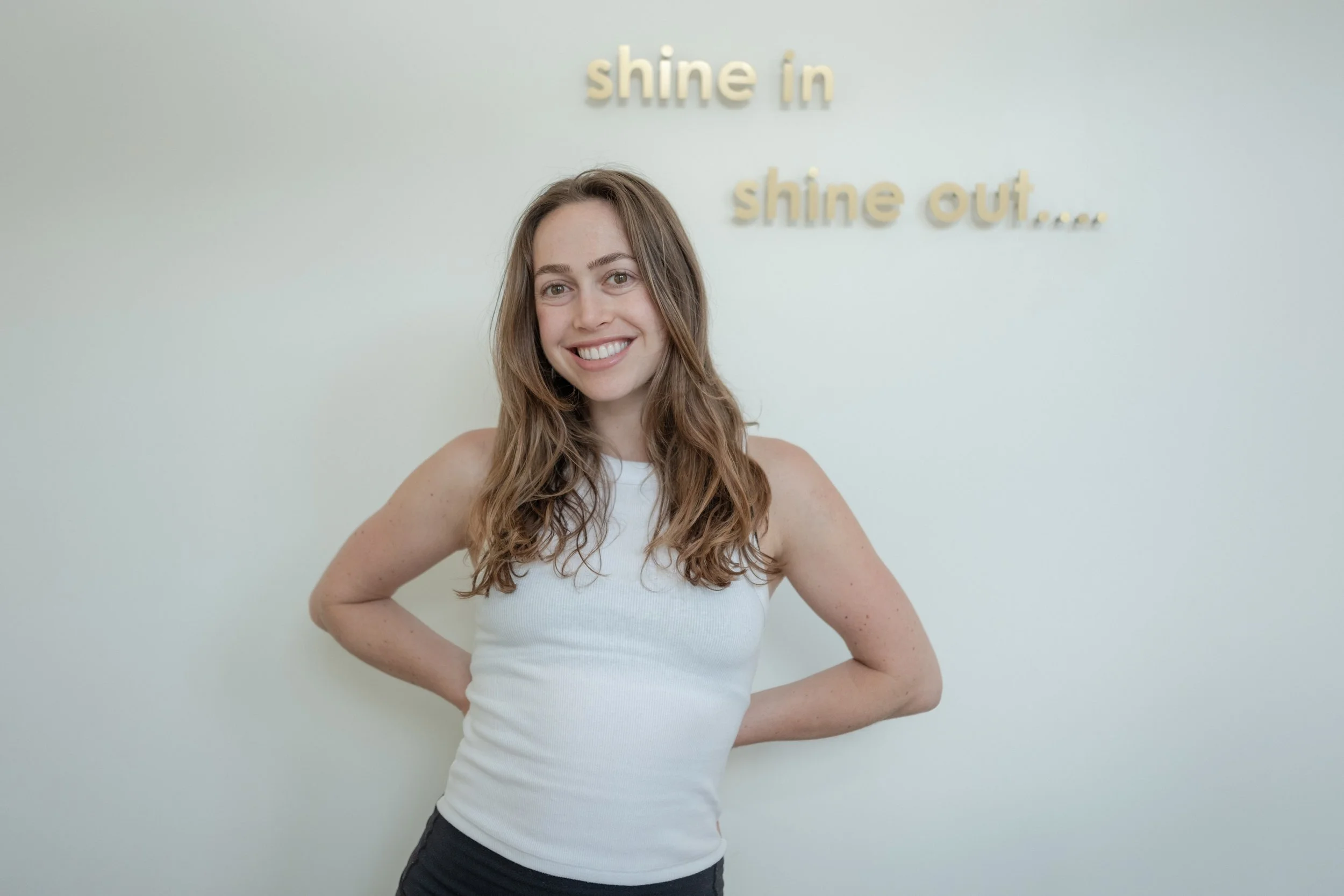 A young woman with long brown hair, smiling, wearing a cream long-sleeve crop top and dark pants, standing against a light-colored wall.