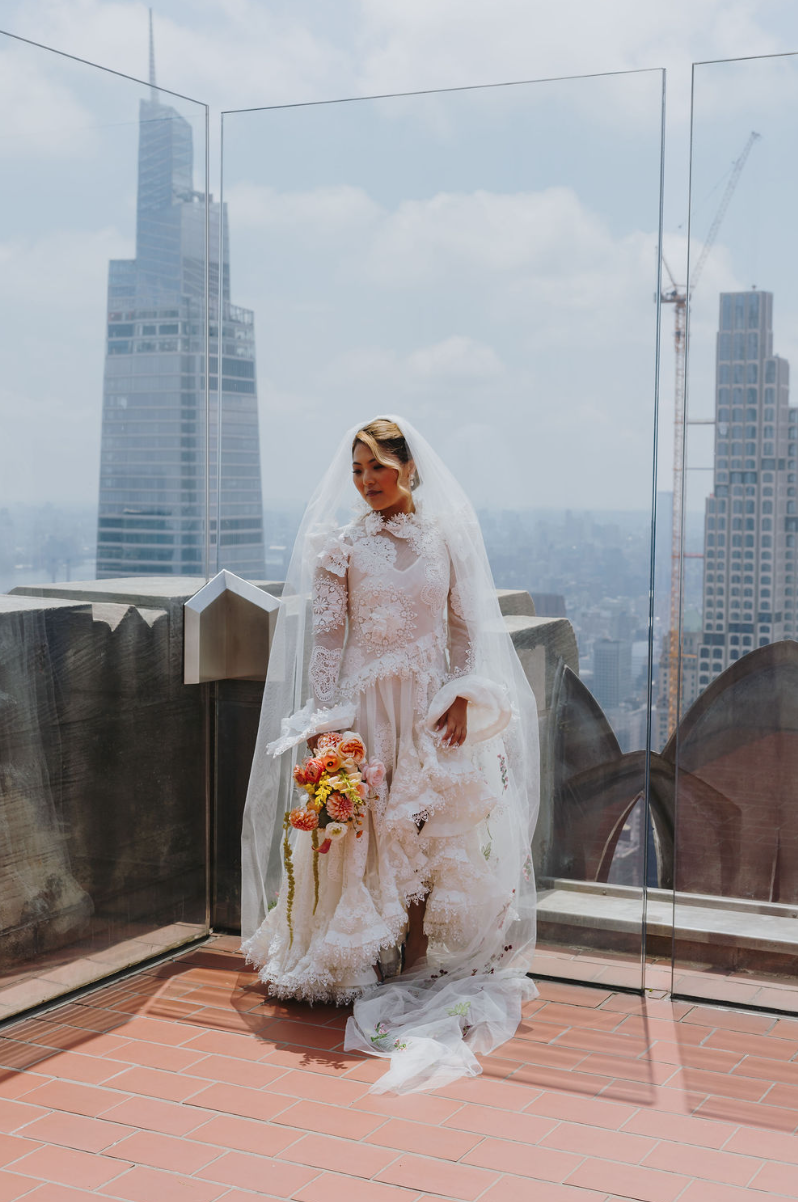 A bride in a white lace wedding dress standing on a rooftop terrace with a cityscape backdrop, holding a bouquet of flowers.