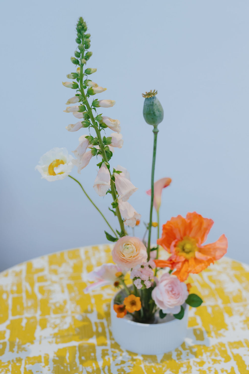 A white vase containing a variety of flowers including a tall lupine, poppies, ranunculus, and other blooms, placed on a yellow patterned cloth.