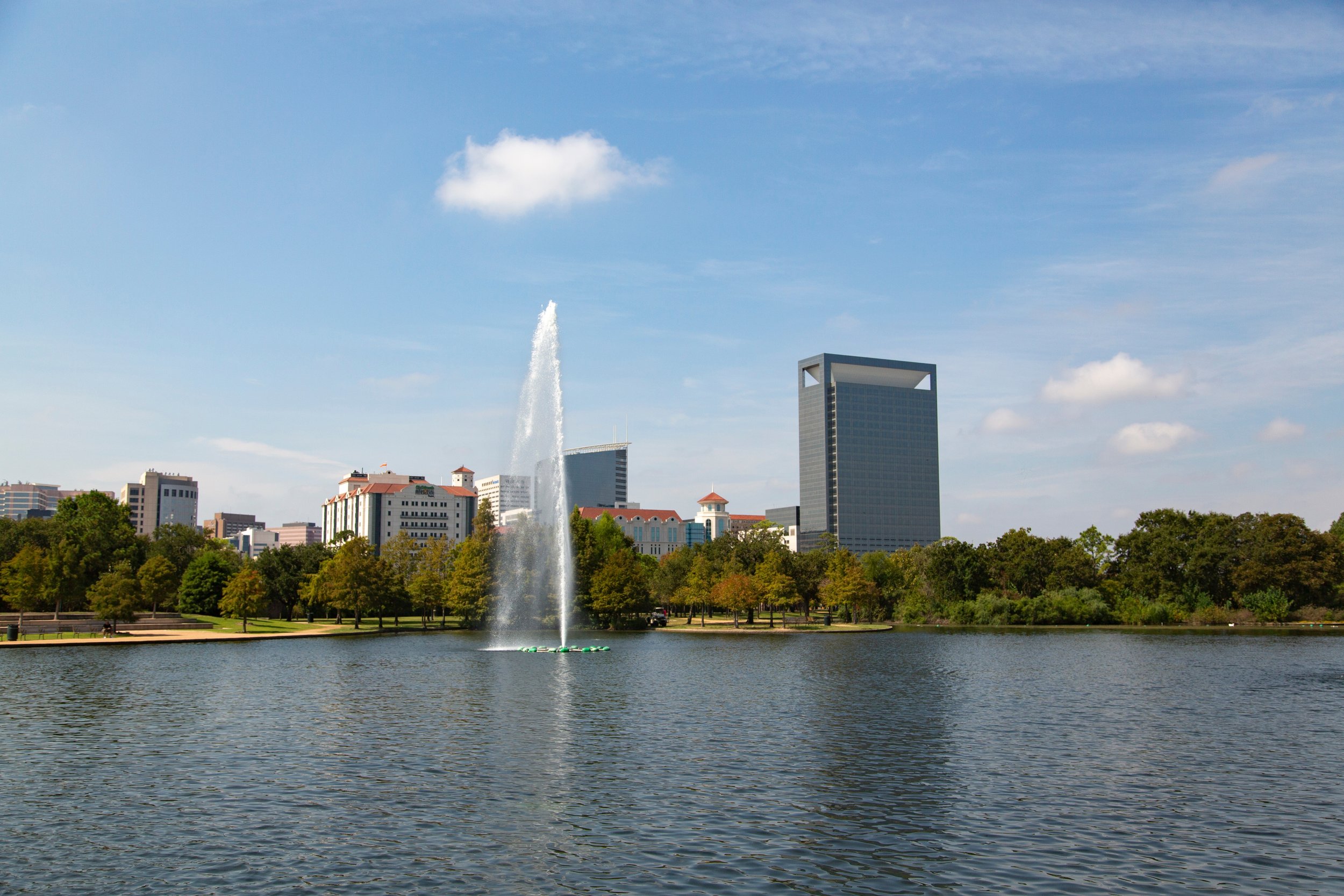 Fountain at Hermann Park's McGovern Lake in Houston, Texas, with trees and Texas Medical Center buildings in the background.