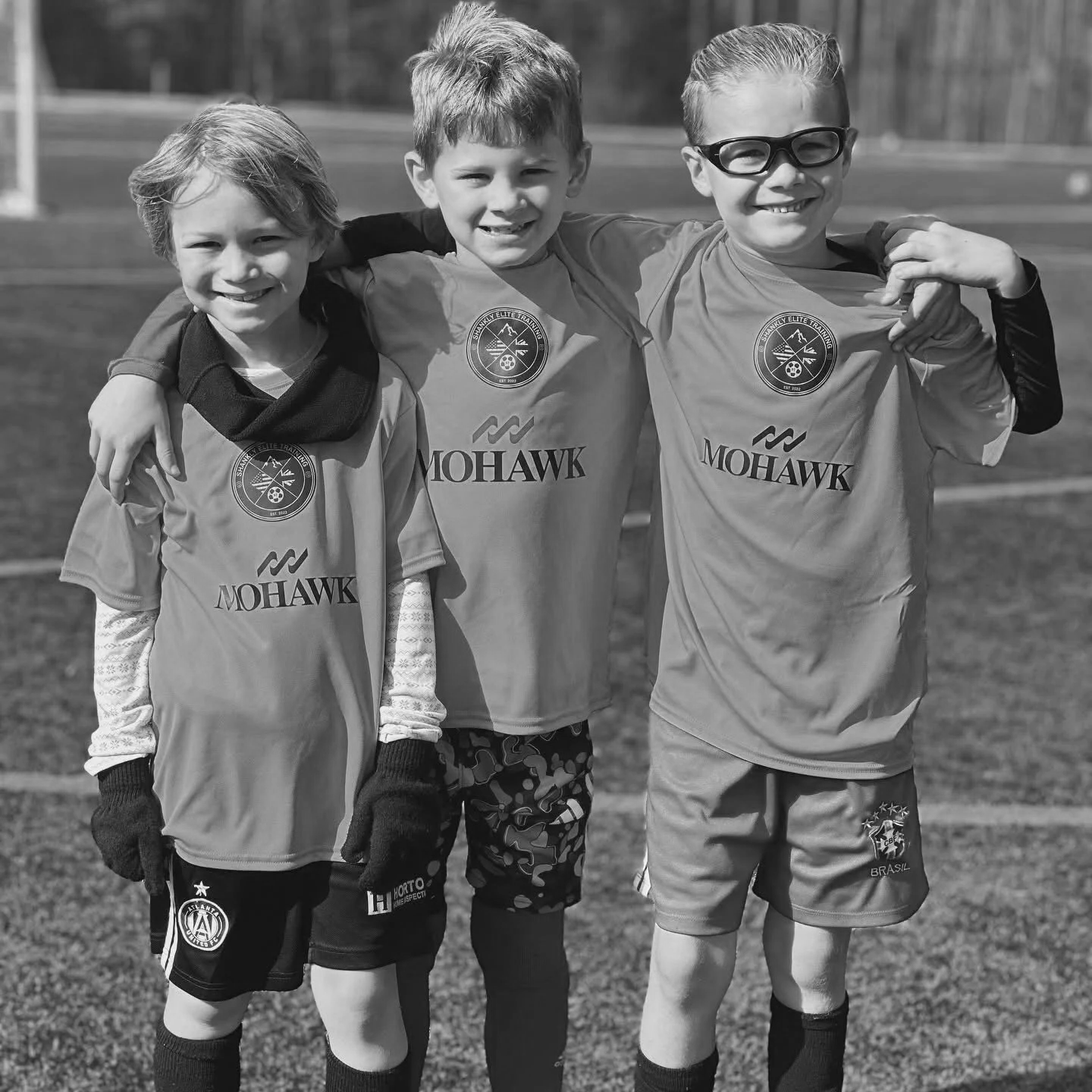 Three young boys standing on a soccer field with their arms around each other's shoulders, wearing soccer jerseys, smiling at the camera.