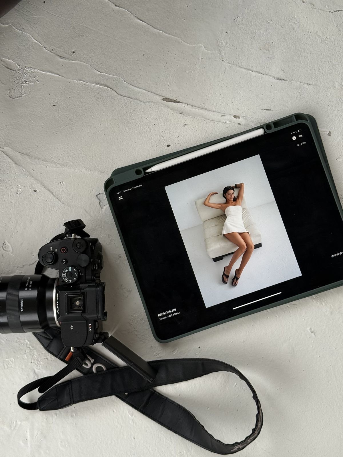 Photo of a DSLR camera and a digital tablet with a photo of a woman in a white dress lying on a chair, on a white textured surface.