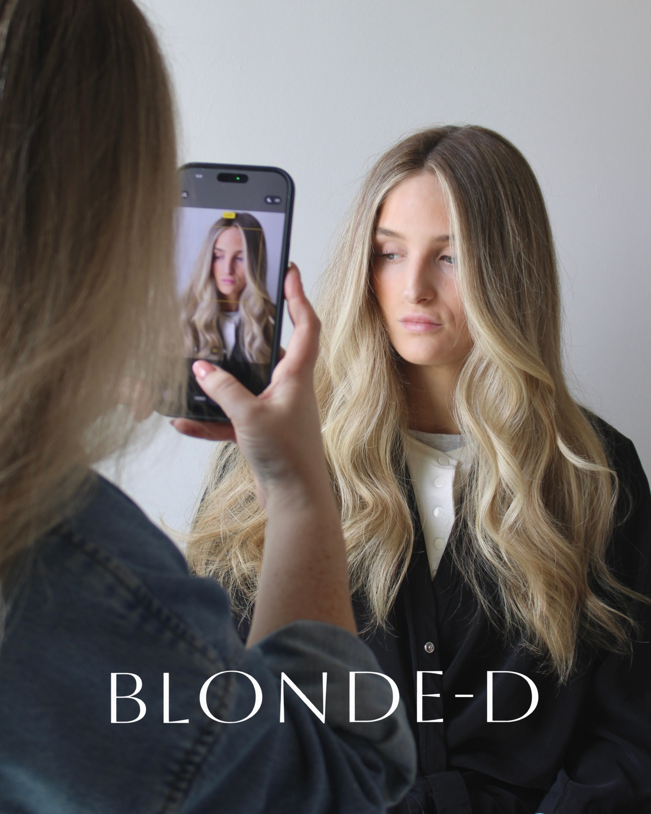 A woman with blonde hair poses for a mirror selfie while another person captures the photo with a phone. The woman’s hair is styled in loose waves and she is wearing a black blazer over a white top. The image is focused on her face and hair, with part of her face showing a neutral expression. The person taking the photo has long, light brown or blonde hair and is wearing a dark sweater.