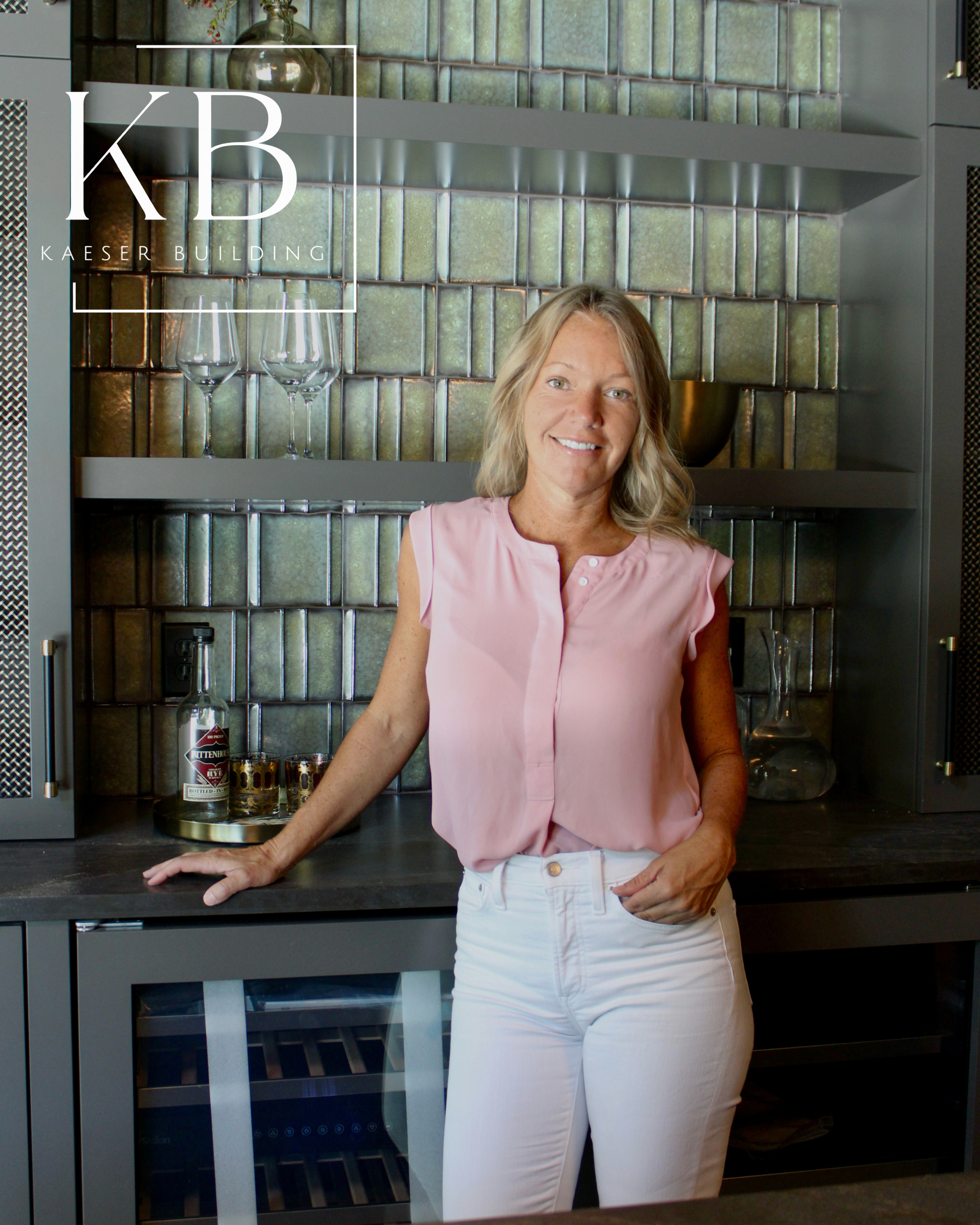 A woman standing in front of a kitchen backsplash with her right arm resting on a countertop, wearing a light pink sleeveless blouse and white pants, smiling at the camera.
