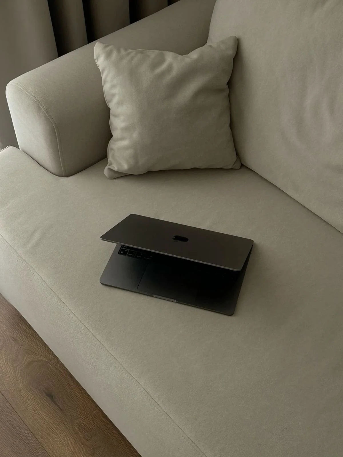A beige couch with a matching pillow, a closed silver laptop, and a glimpse of hardwood floor.