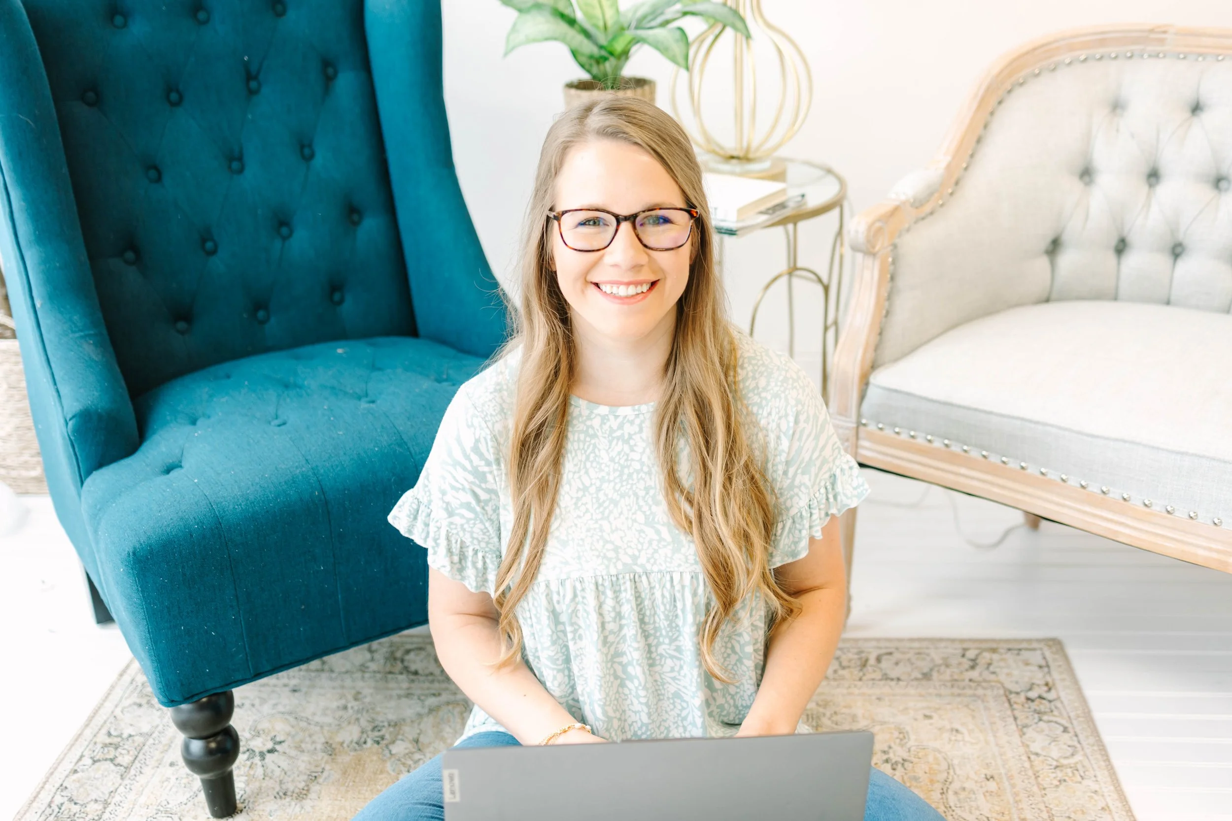 A smiling woman with long blonde hair and glasses sitting on the floor with a laptop in front of her in a bright, cozy living room with a teal armchair, a beige loveseat, a side table, a potted plant, and a rug.