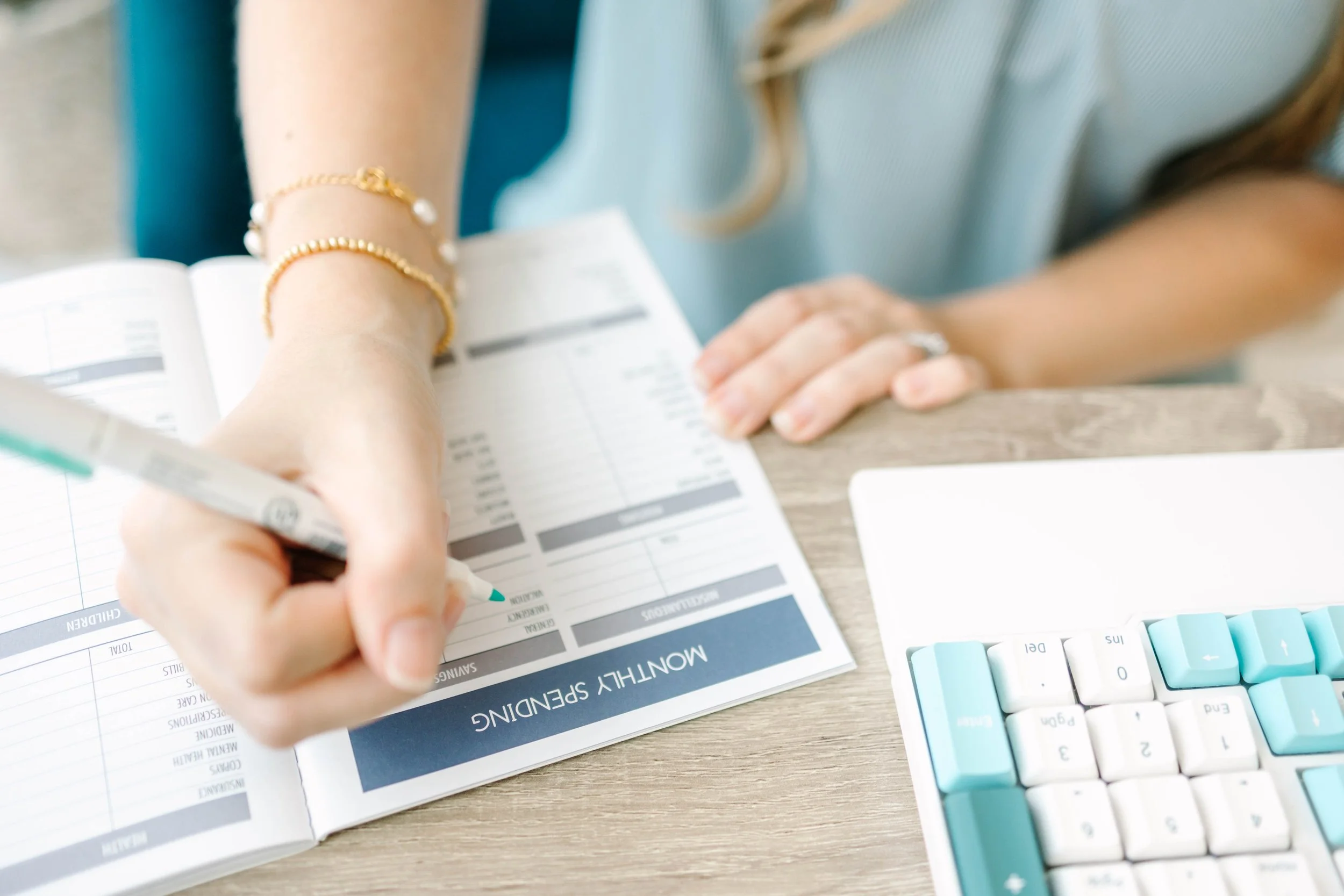 A person filling out a monthly spending planner in a notebook at a desk. The person is wearing a ring and a gold bracelet, with a keyboard visible nearby.