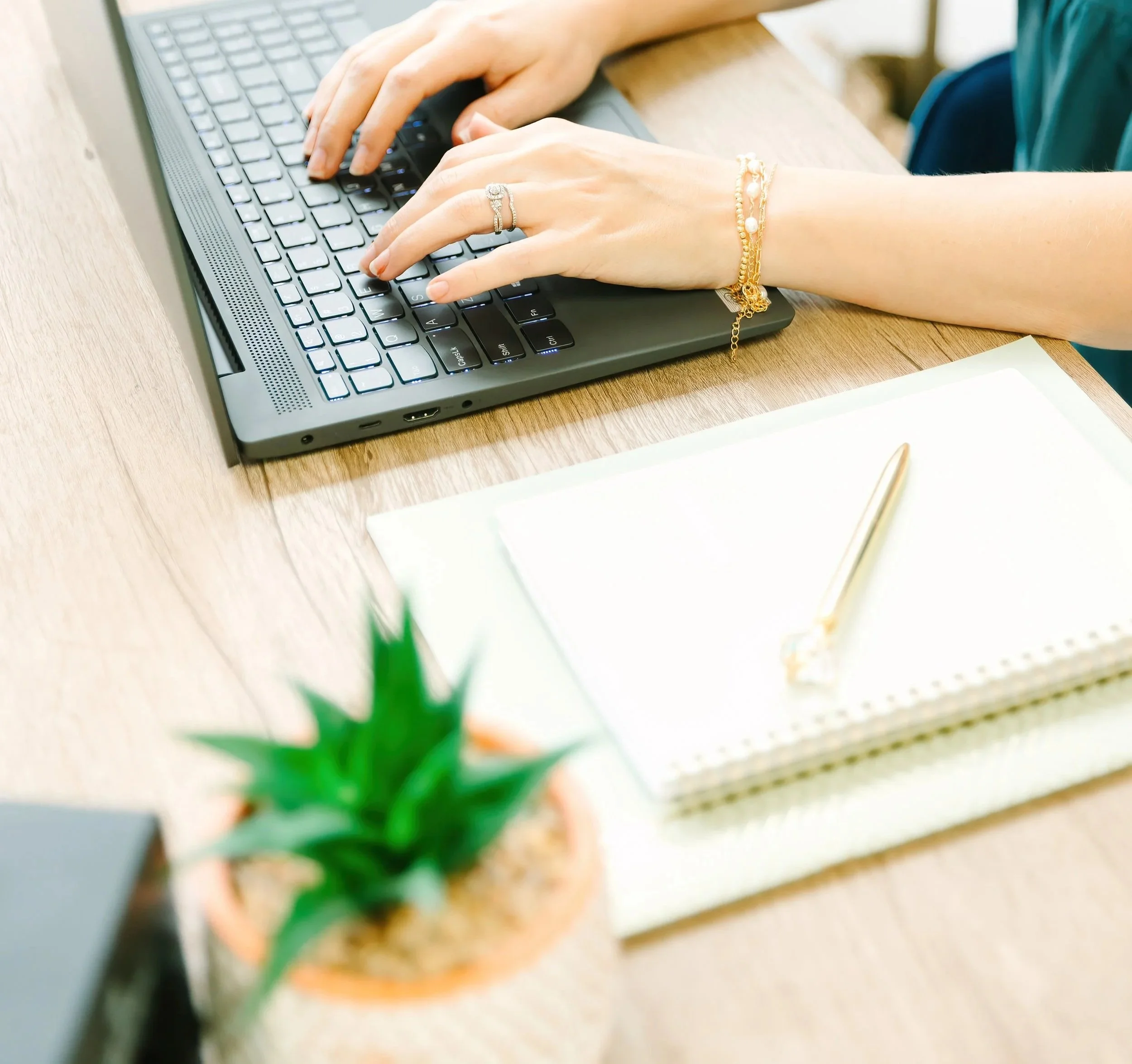 Person typing on a laptop keyboard on a wooden desk, with a notebook and pen nearby, and a small potted cactus in the foreground.