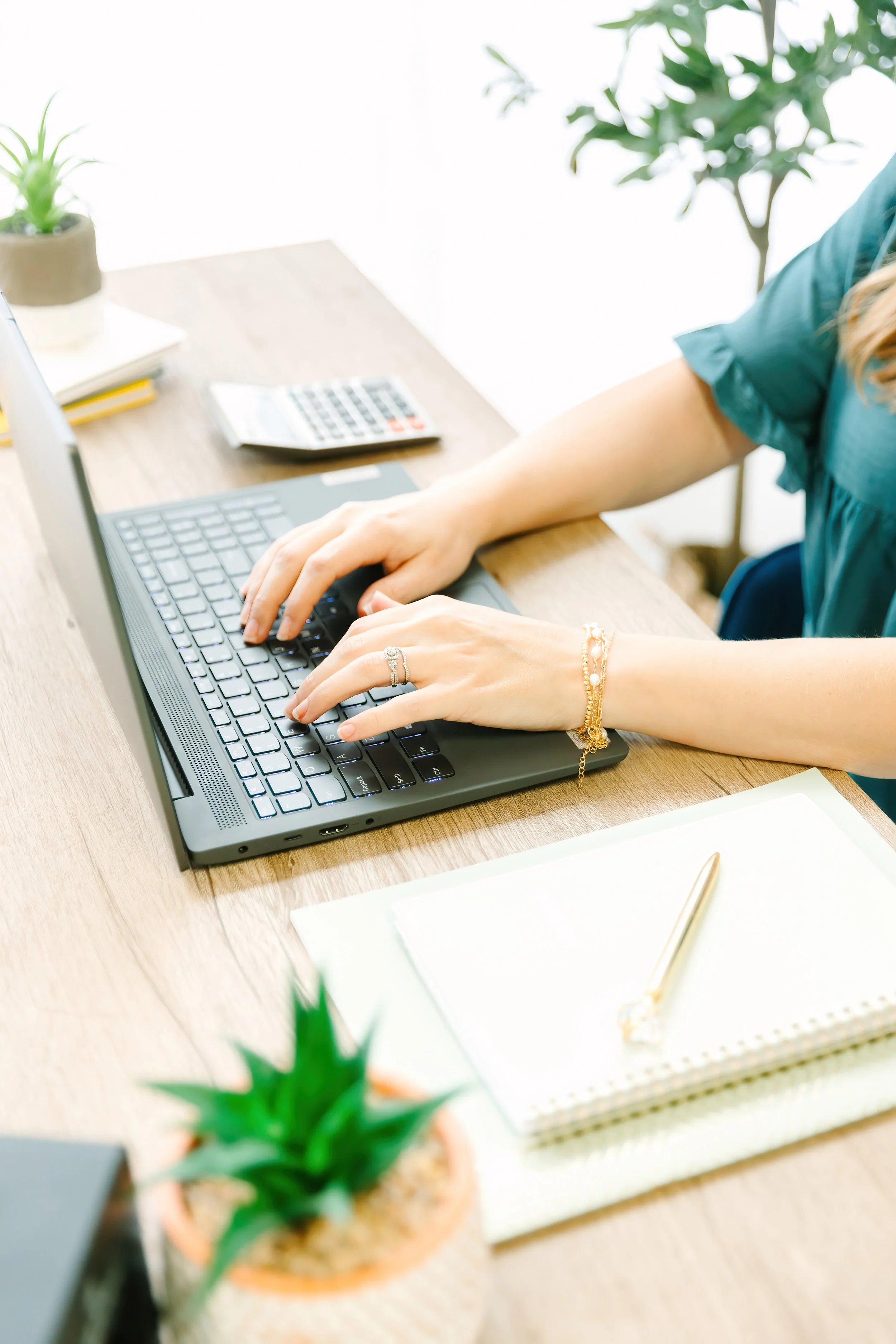 Person typing on a laptop at a wooden desk with potted plants and a calculator.