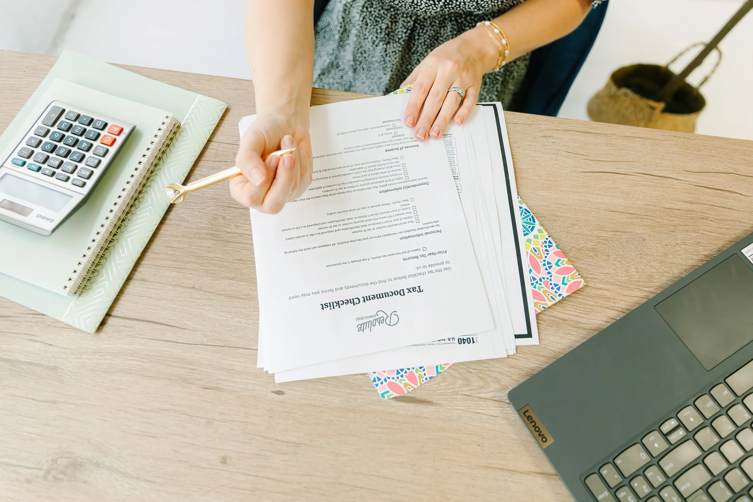 Person reviewing financial documents at a desk, with a calculator, notebook, colorful folder, and a laptop.