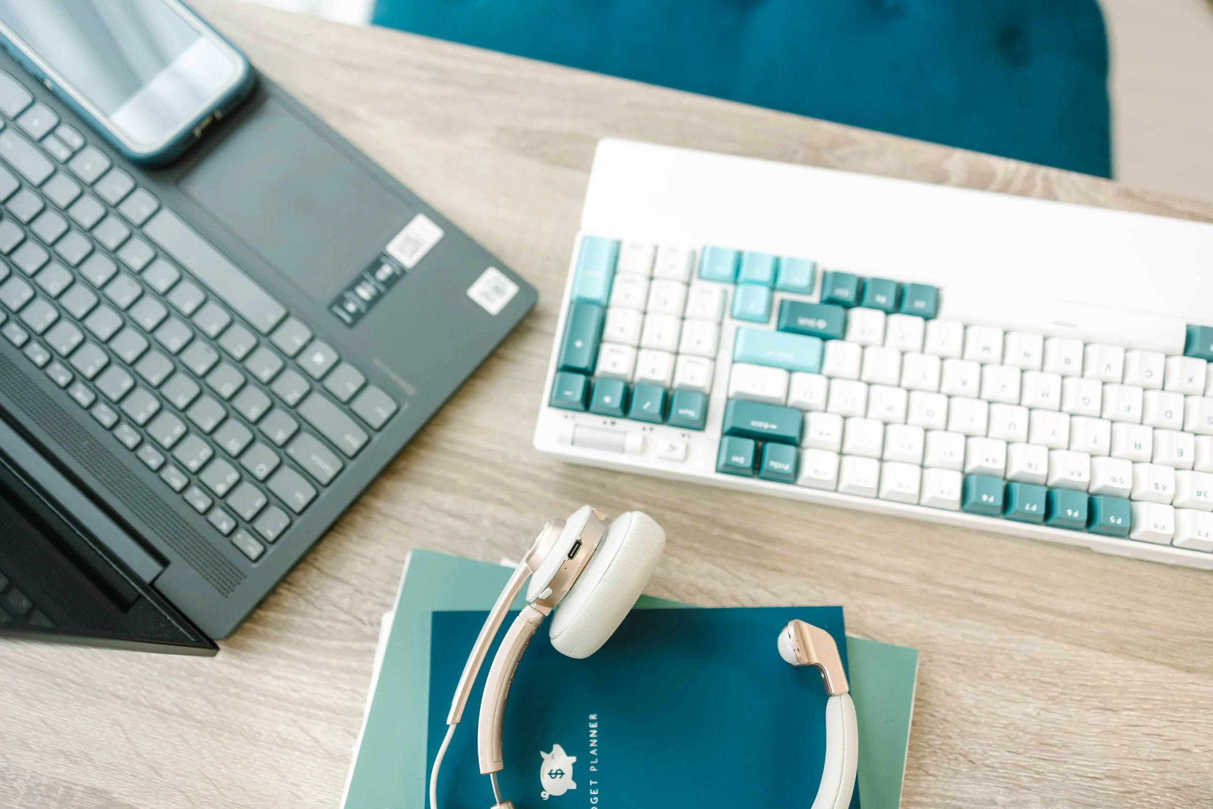 Desk with a laptop, a smartphone, a mechanical keyboard with teal and white keys, a pair of white headphones on a blue planner, and some papers on a wooden surface.