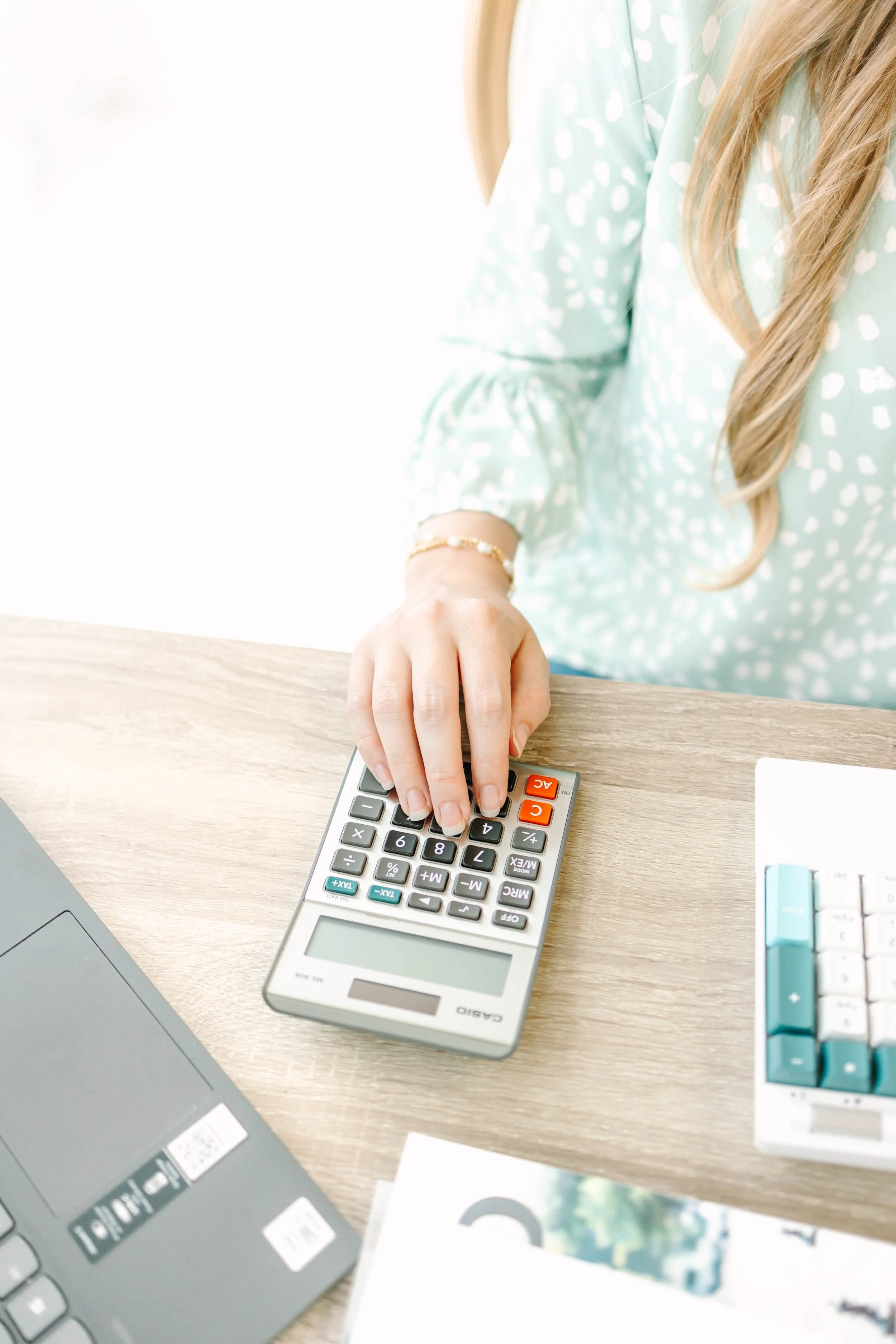 A woman in a light blue patterned blouse using a calculator on a wooden desk, with a laptop and some papers nearby.