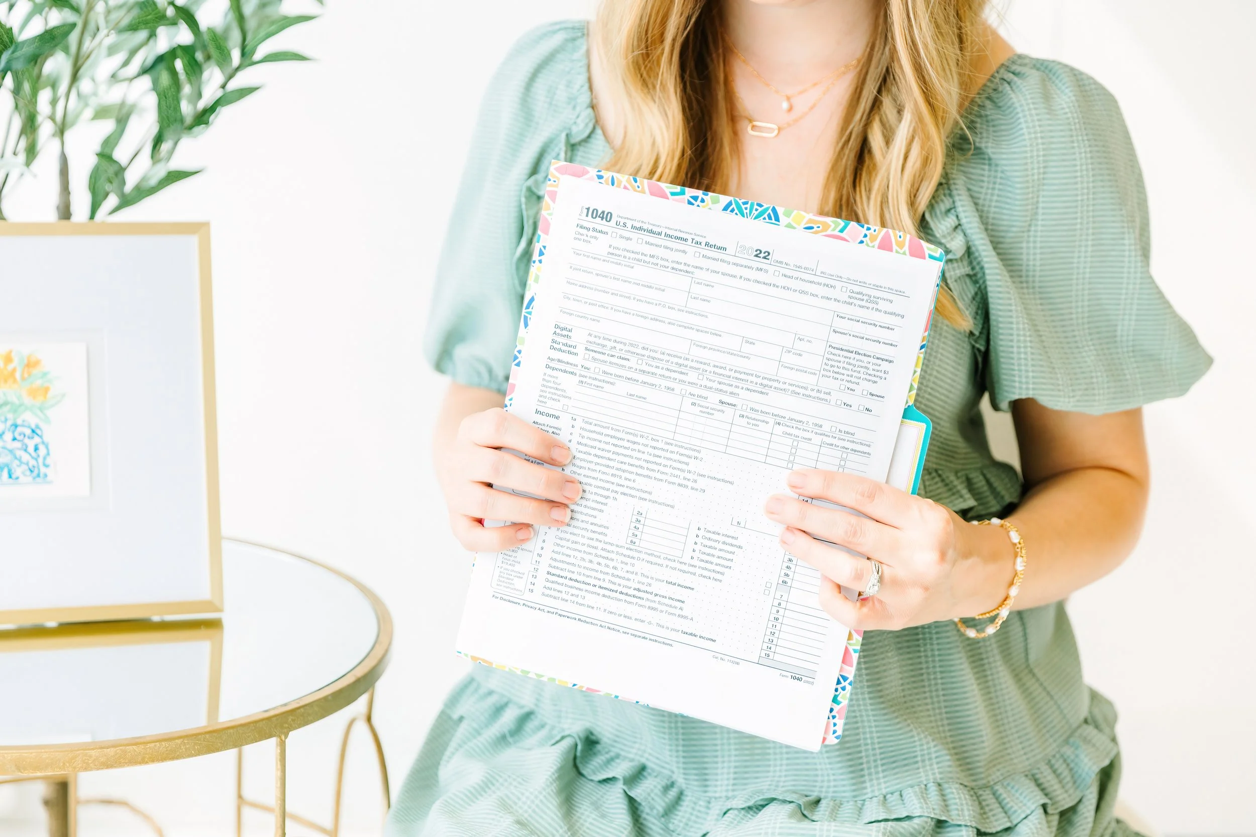 A woman holding a completed United States tax return form. She wears a light green dress, and is seated next to a small round table with a framed piece of artwork and a plant in the background.