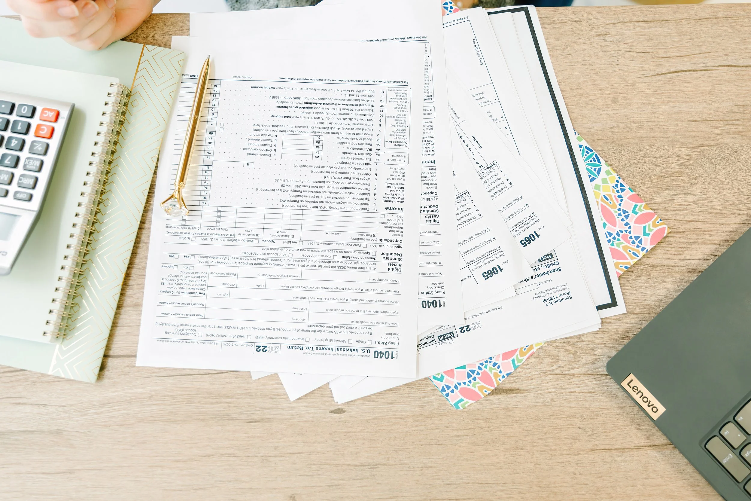 A cluttered desk with papers, a golden pen, a calculator, and a laptop.
