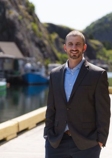 Eddie Sheerr in a suit standing outdoors near a marina with boats and rocky hills in the background.