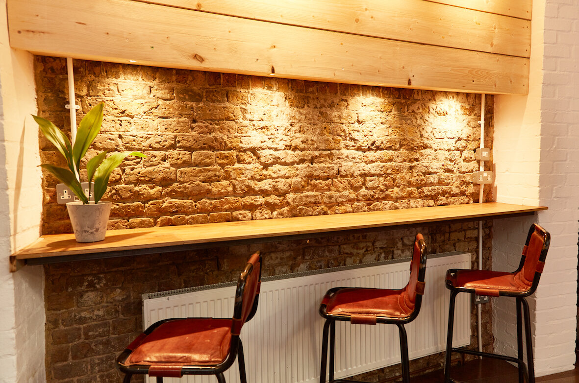 A wooden counter with three chairs, exposed brick wall, potted plant, and electrical outlets with power cords.