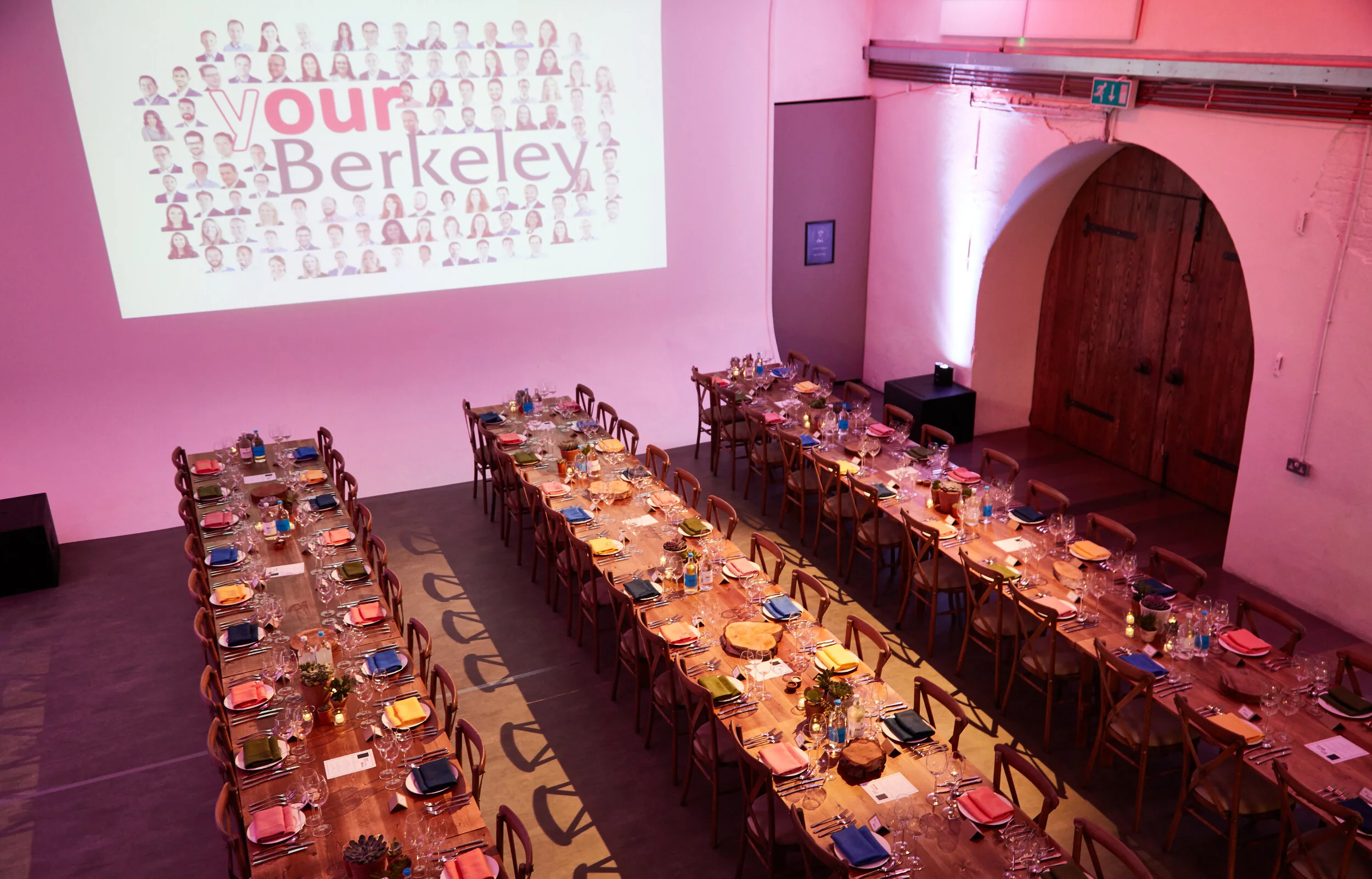 Empty banquet hall with long tables set for a formal event, decorated with colorful napkins, glassware, and small potted plants, illuminated with pink lighting, and a large projector screen displaying 'your Berkeley' with a collage of people's faces.