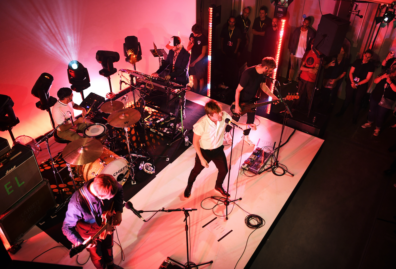 A band performs live on stage with musicians playing drums, guitar, bass, and keyboard, and a singer with a microphone. Audience members watch from the side and in the background with red lighting illuminating the scene.
