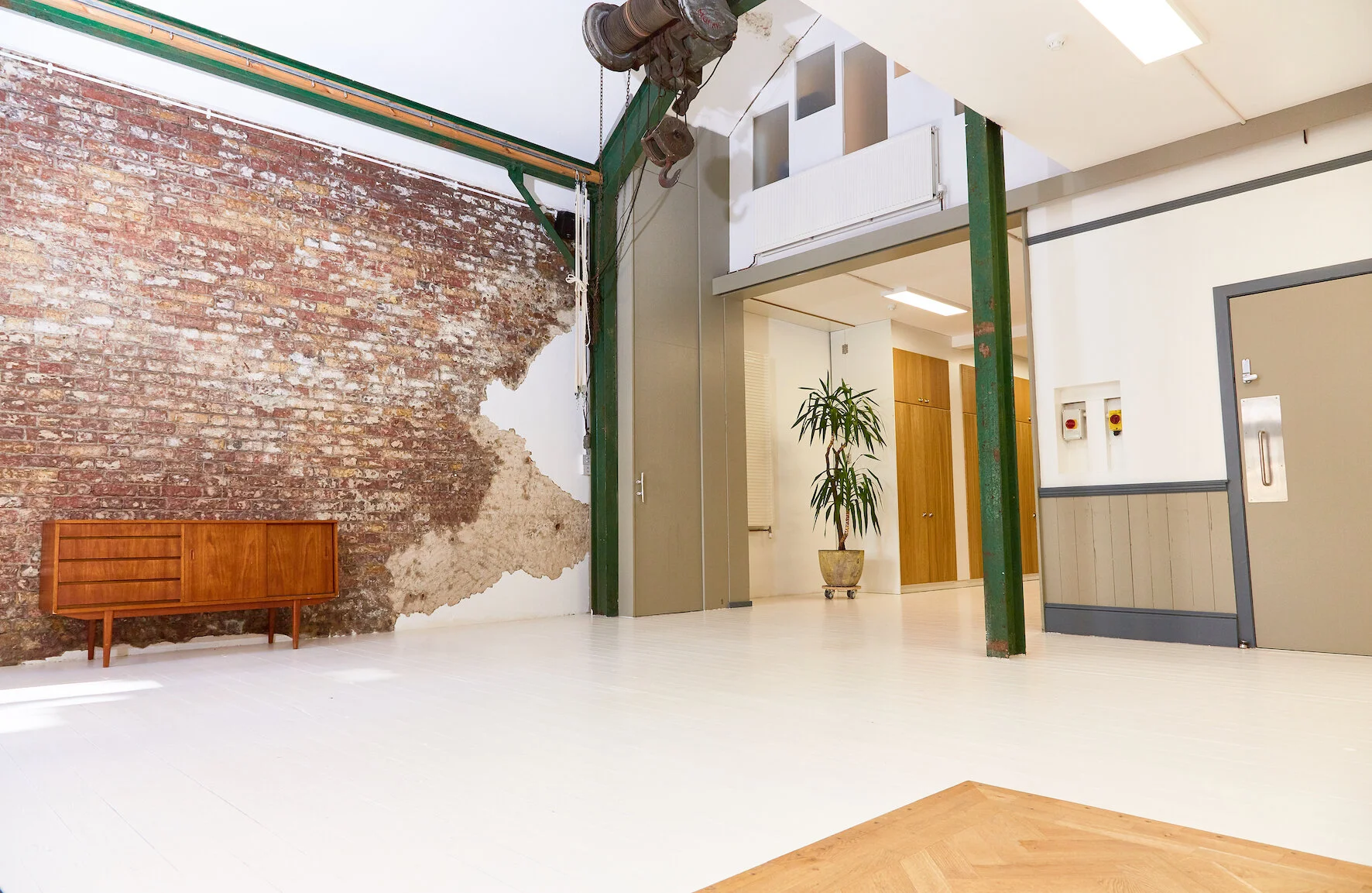 Interior view of an industrial-style room with a partially exposed brick wall, white flooring, and a wooden sideboard. A green metal beam supports the ceiling, and there's a large potted plant near wooden cabinets. The ceiling has skylights and some mechanical equipment hanging.
